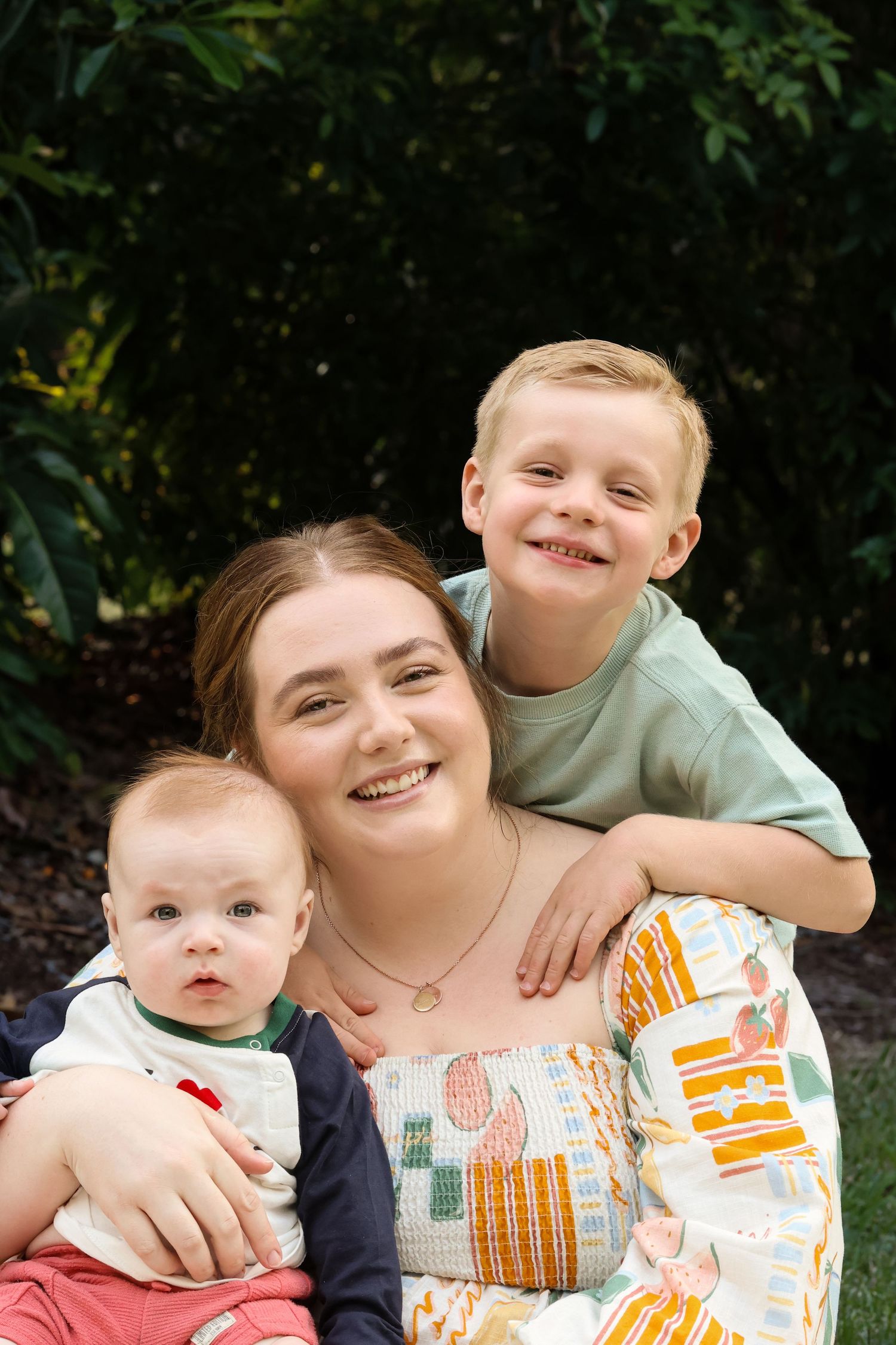 A young woman smiles outdoors holding a baby while an older boy hugs her from behind, surrounded by greenery.