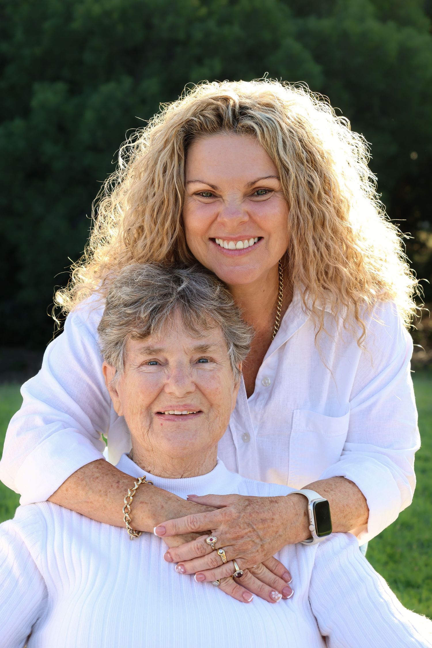 Two women in white shirts smile outdoors, younger woman hugging older woman from behind near green trees.