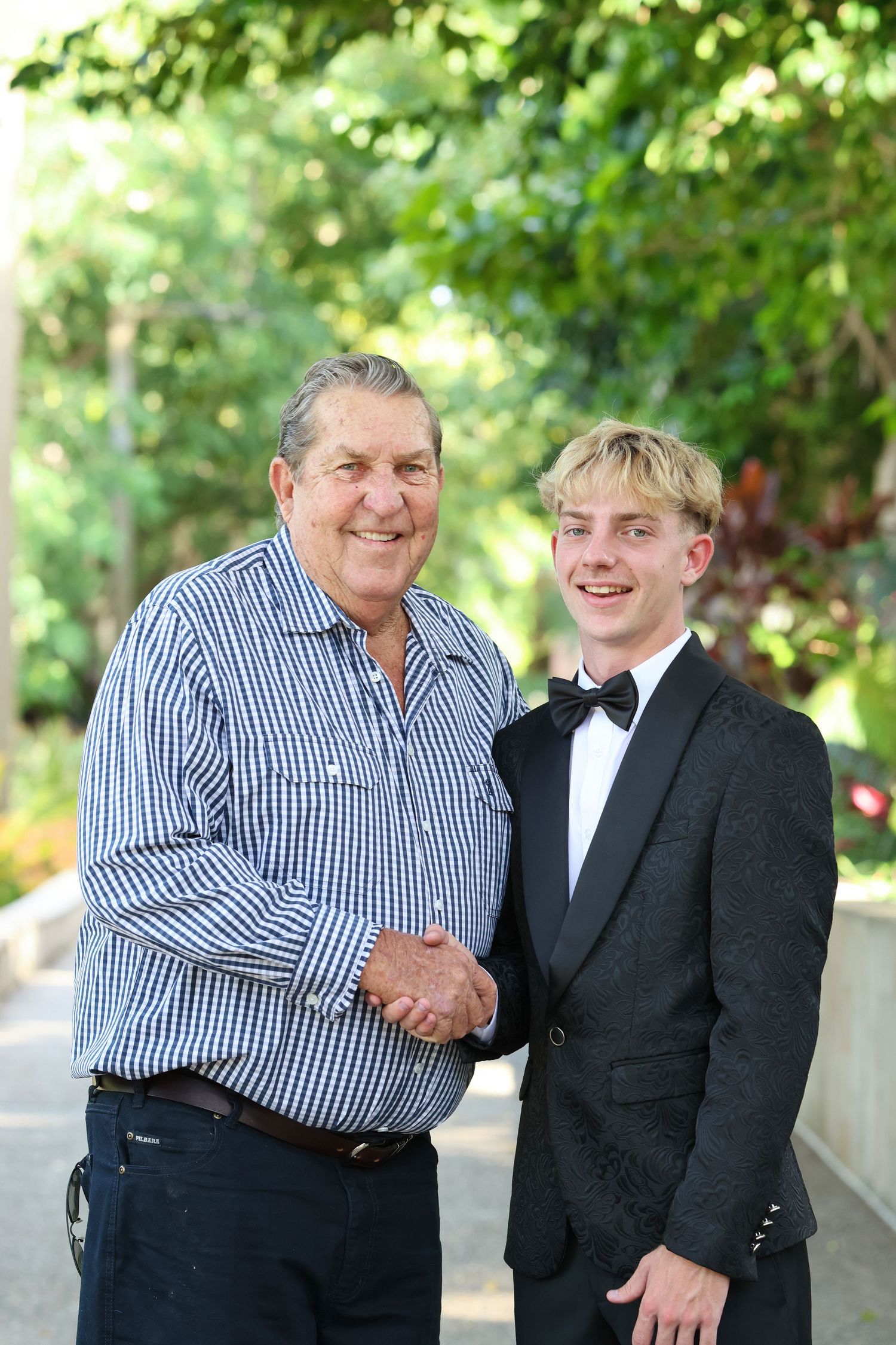 An older man and a teenage boy in a tuxedo shaking hands and smiling outdoors among green trees.