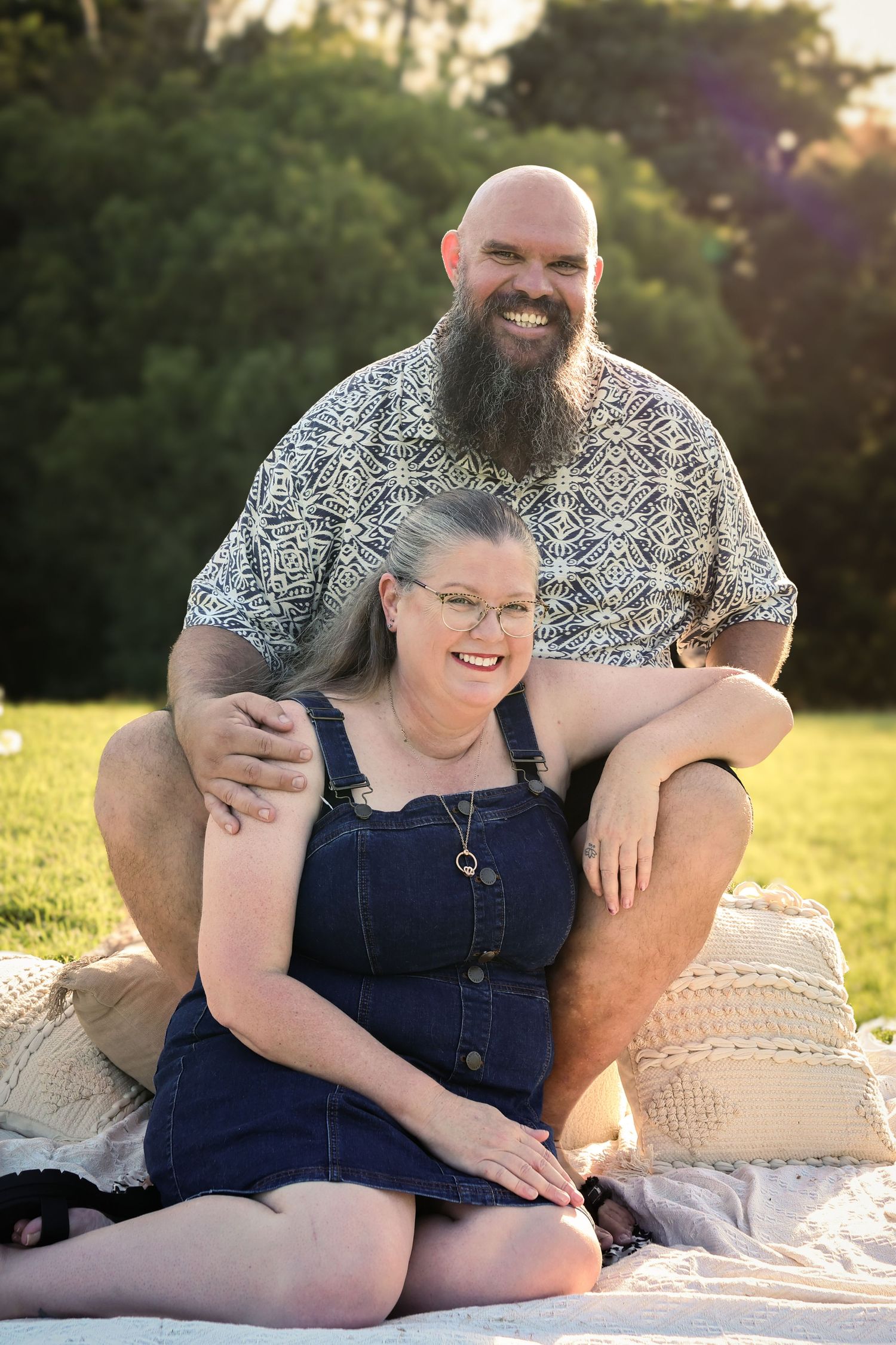 A smiling couple poses outdoors on a blanket, man with beard sits behind woman in denim overalls, green trees background.