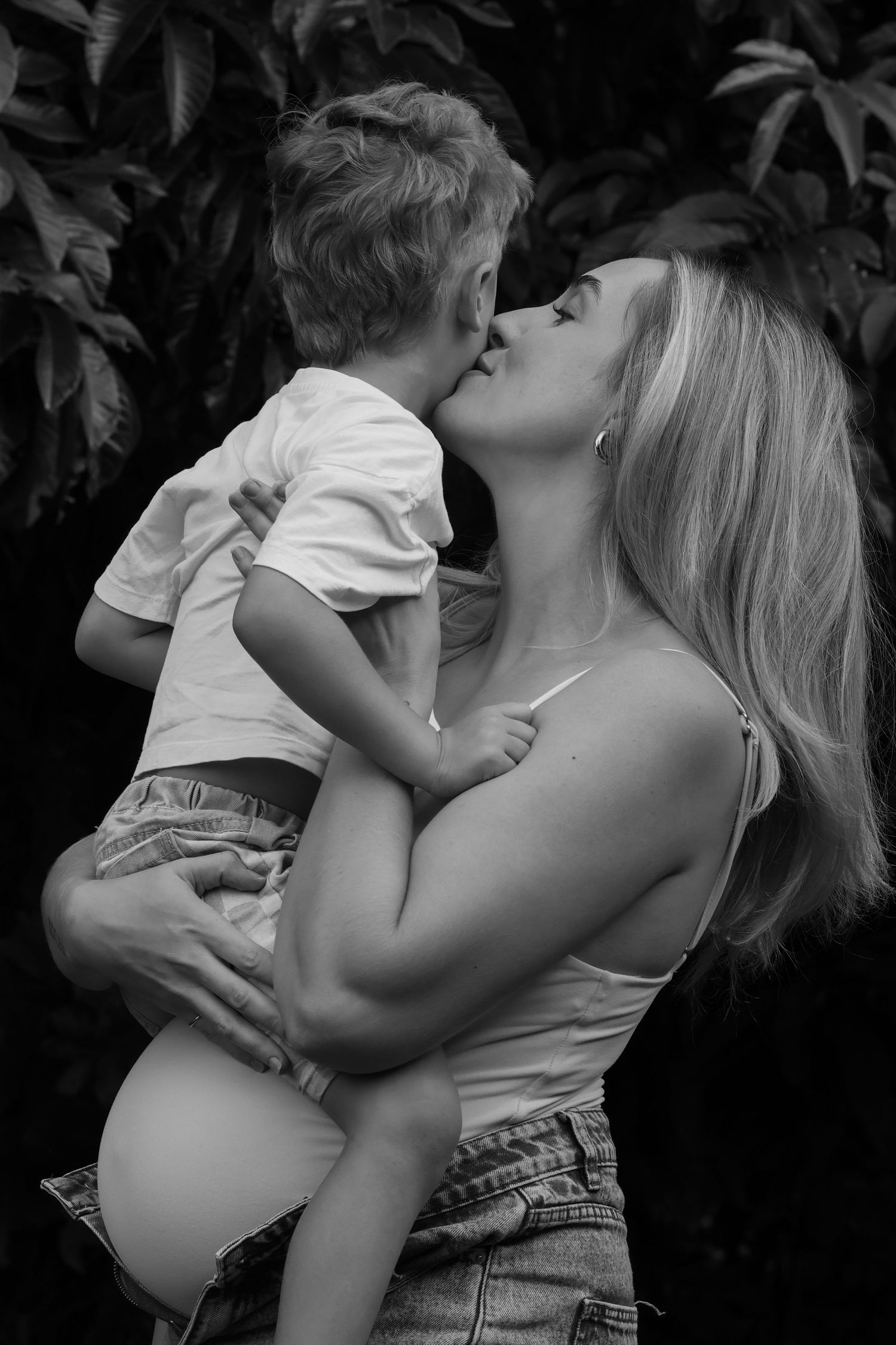 Black and white photo of a blonde woman holding and kissing a young child outdoors near lush green foliage.