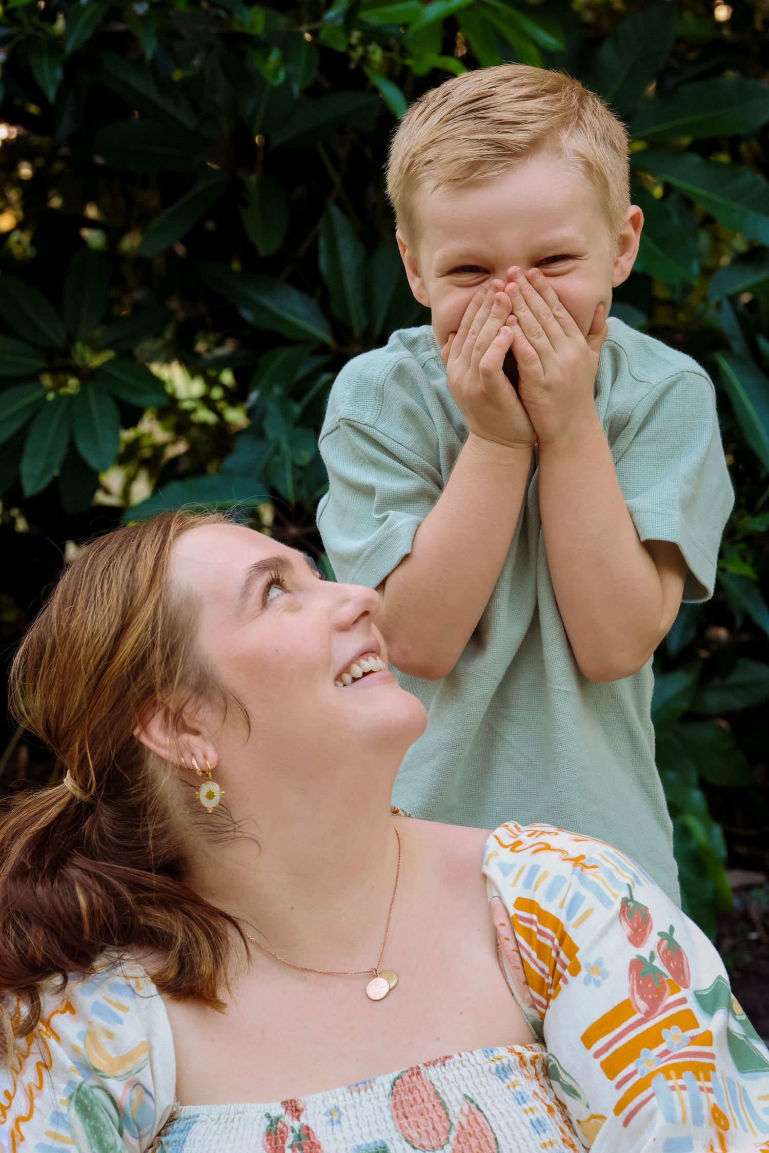 A joyful mother looks up smiling at her laughing young blond boy covering his mouth outdoors.