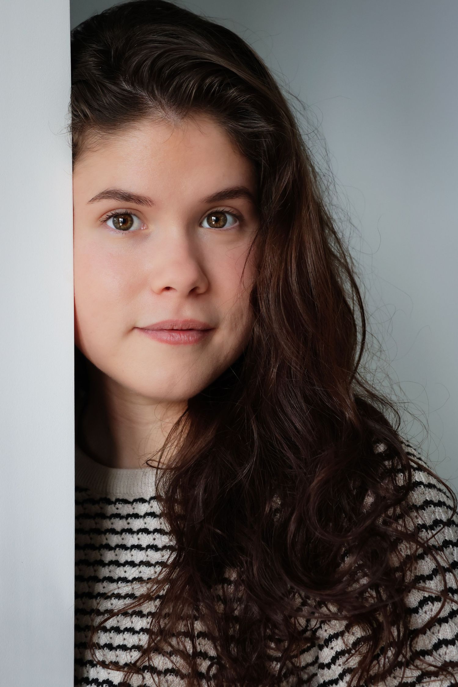 Young woman with long brown wavy hair leans against a white wall, wearing a striped sweater, gazing at camera.