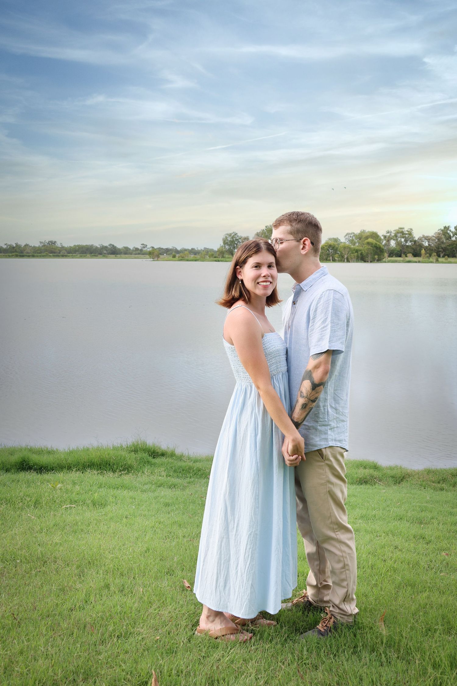 A couple shares a tender moment by a serene lake at sunset, standing on green grass in casual summer attire.
