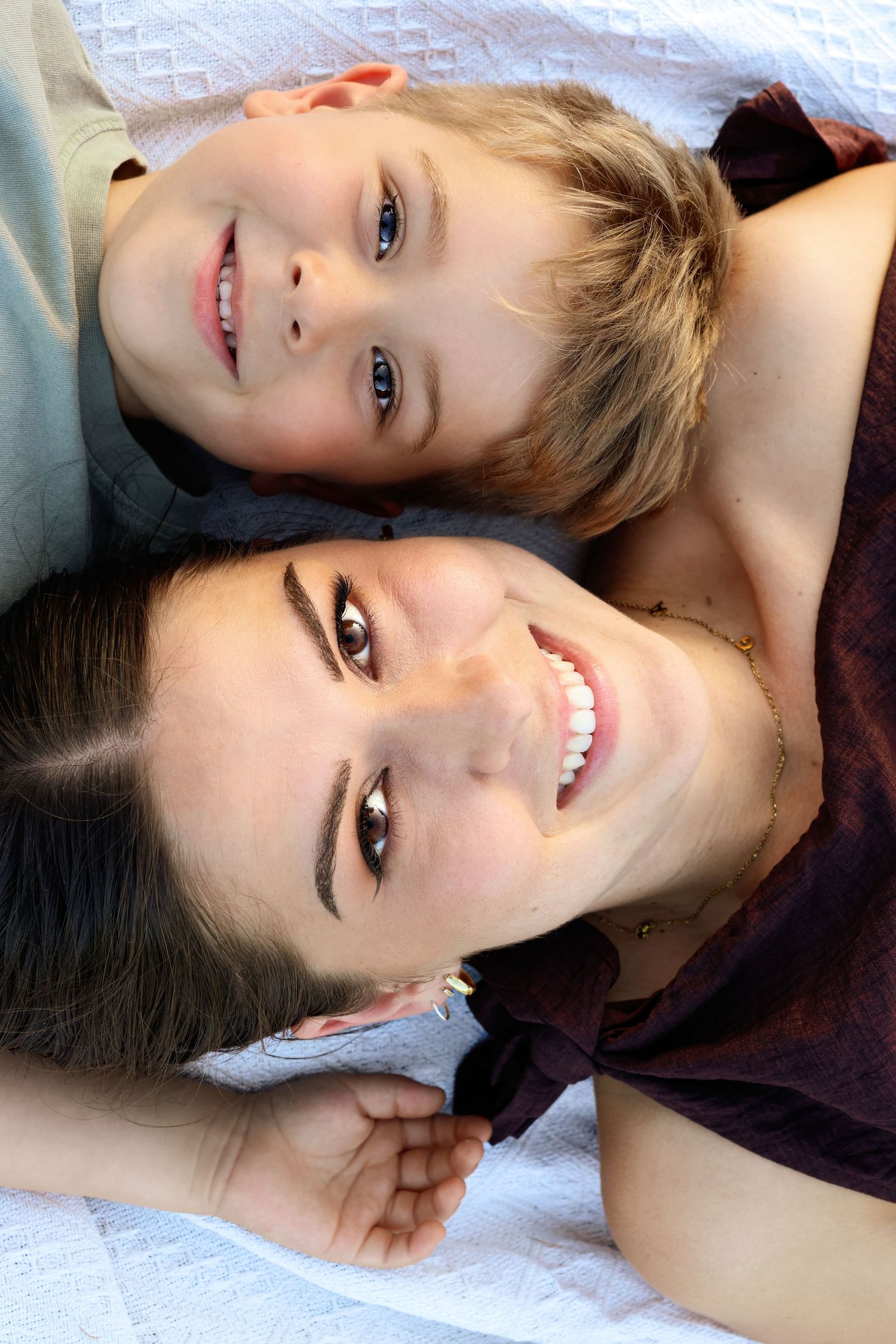 A smiling mother and young son lying together on a white blanket, looking up at the camera with joyful expressions.