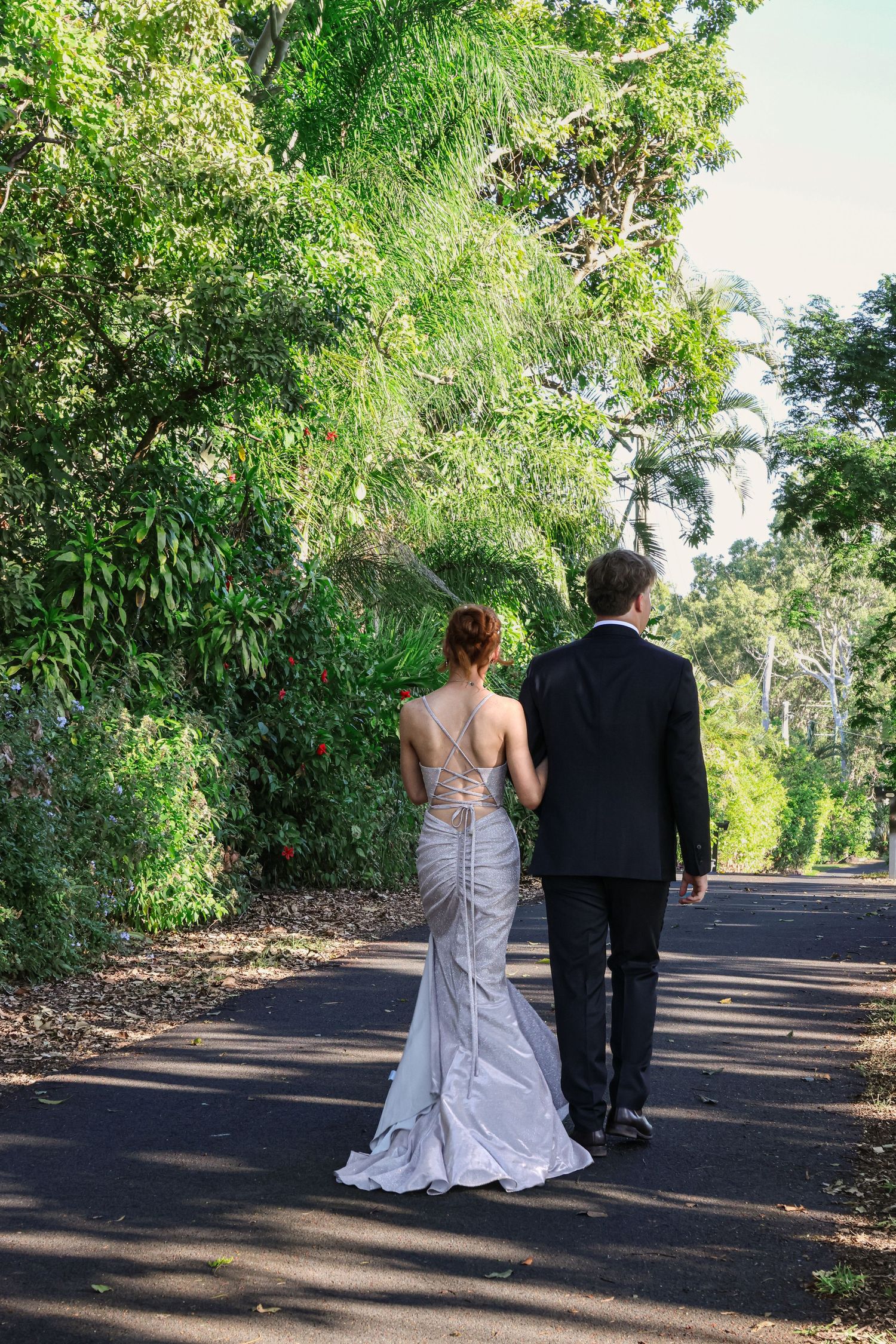 Bride and groom walking hand in hand down a tree-lined path, bride in elegant backless gown, groom in black suit.