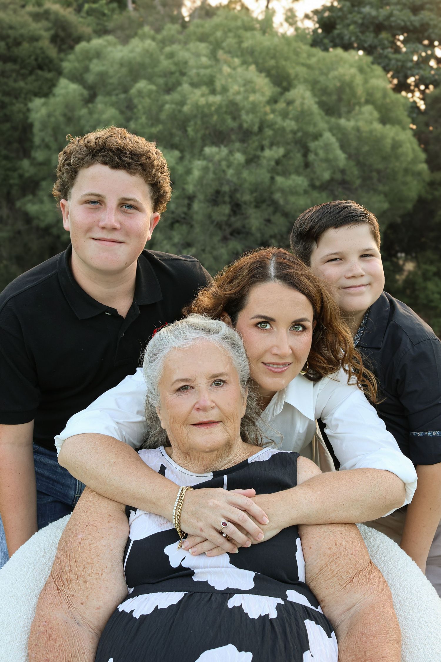 Four generations of family posing outdoors, elderly woman seated in front with younger woman and two teenage boys behind her.