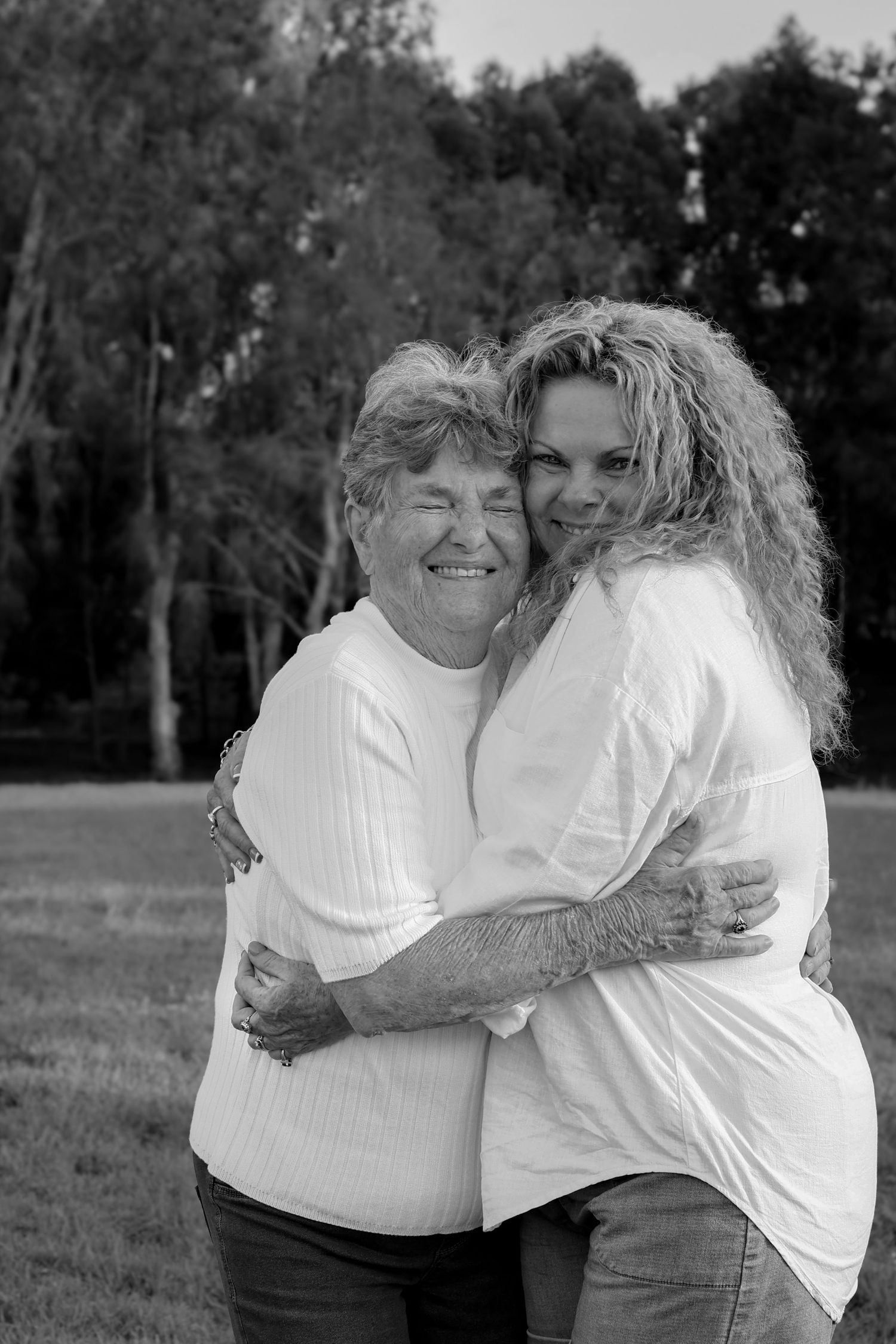 Two women hugging and smiling outdoors in black and white photo, wearing white tops in a park setting.