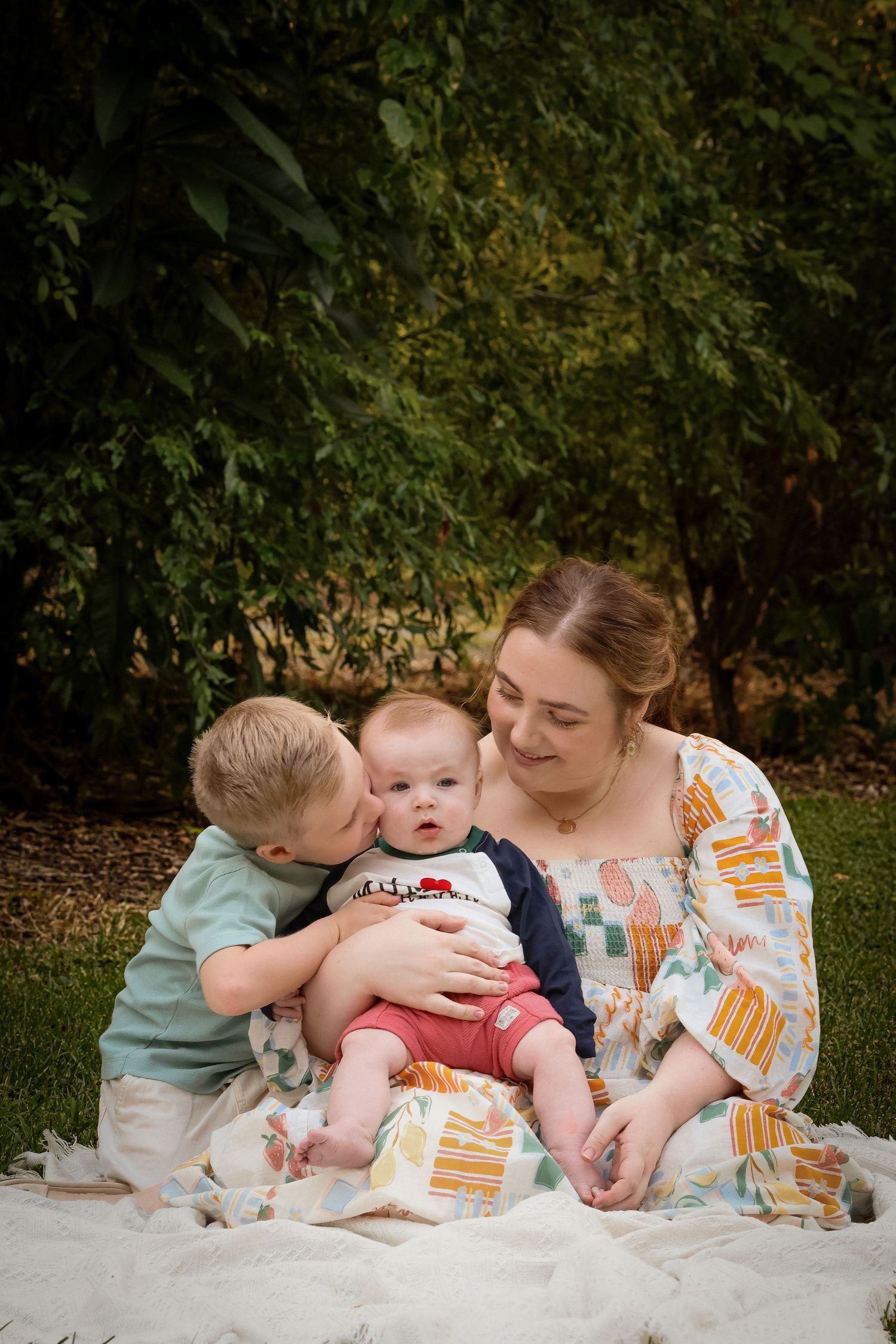 A mother sits on a blanket outdoors with her two young children, smiling warmly as her toddler kisses the baby.