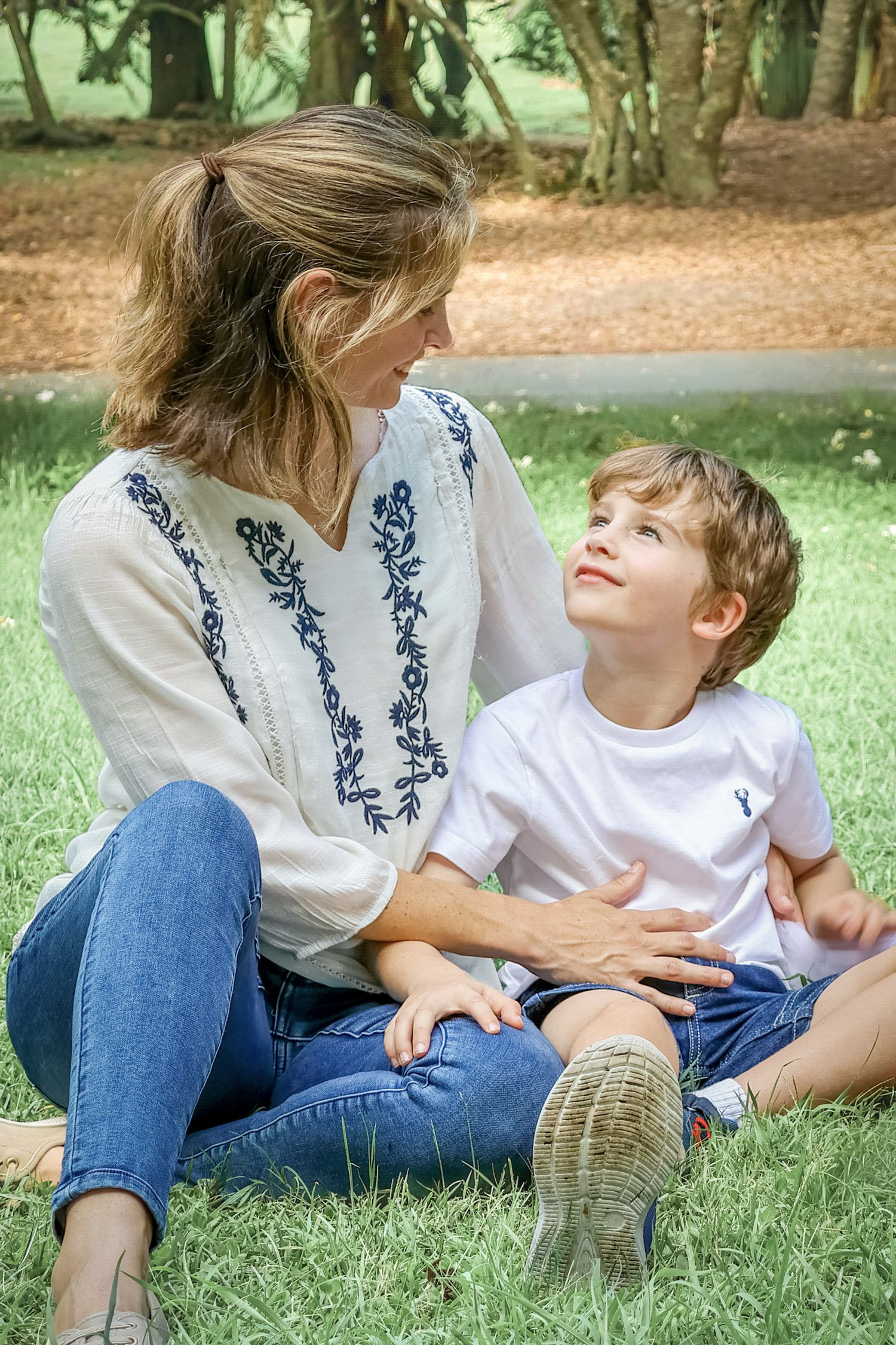 A woman and young boy sit together on green grass in a park, smiling at each other warmly.