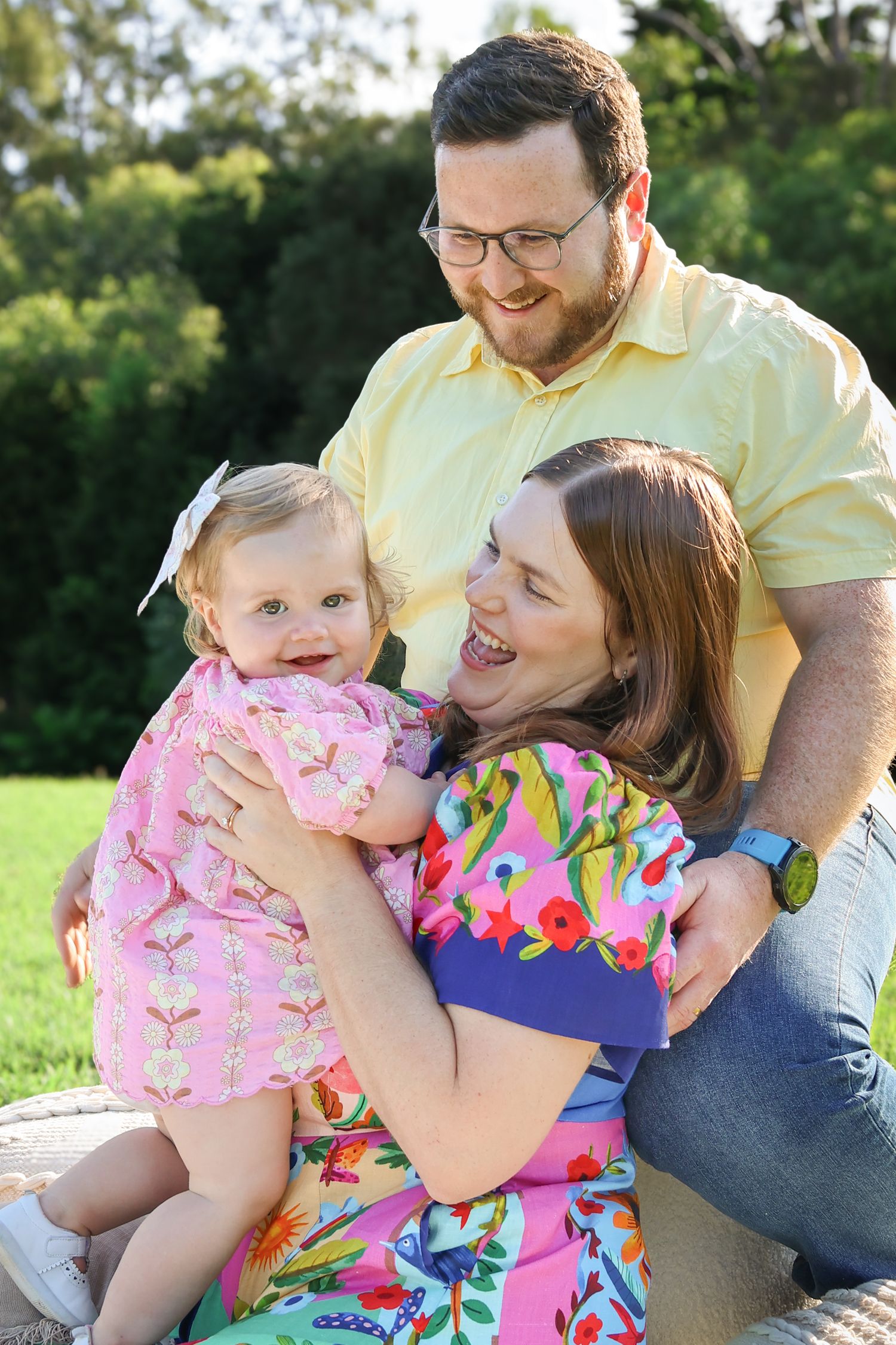 Happy family with parents and baby girl laughing outdoors in a lush green garden on a sunny day.