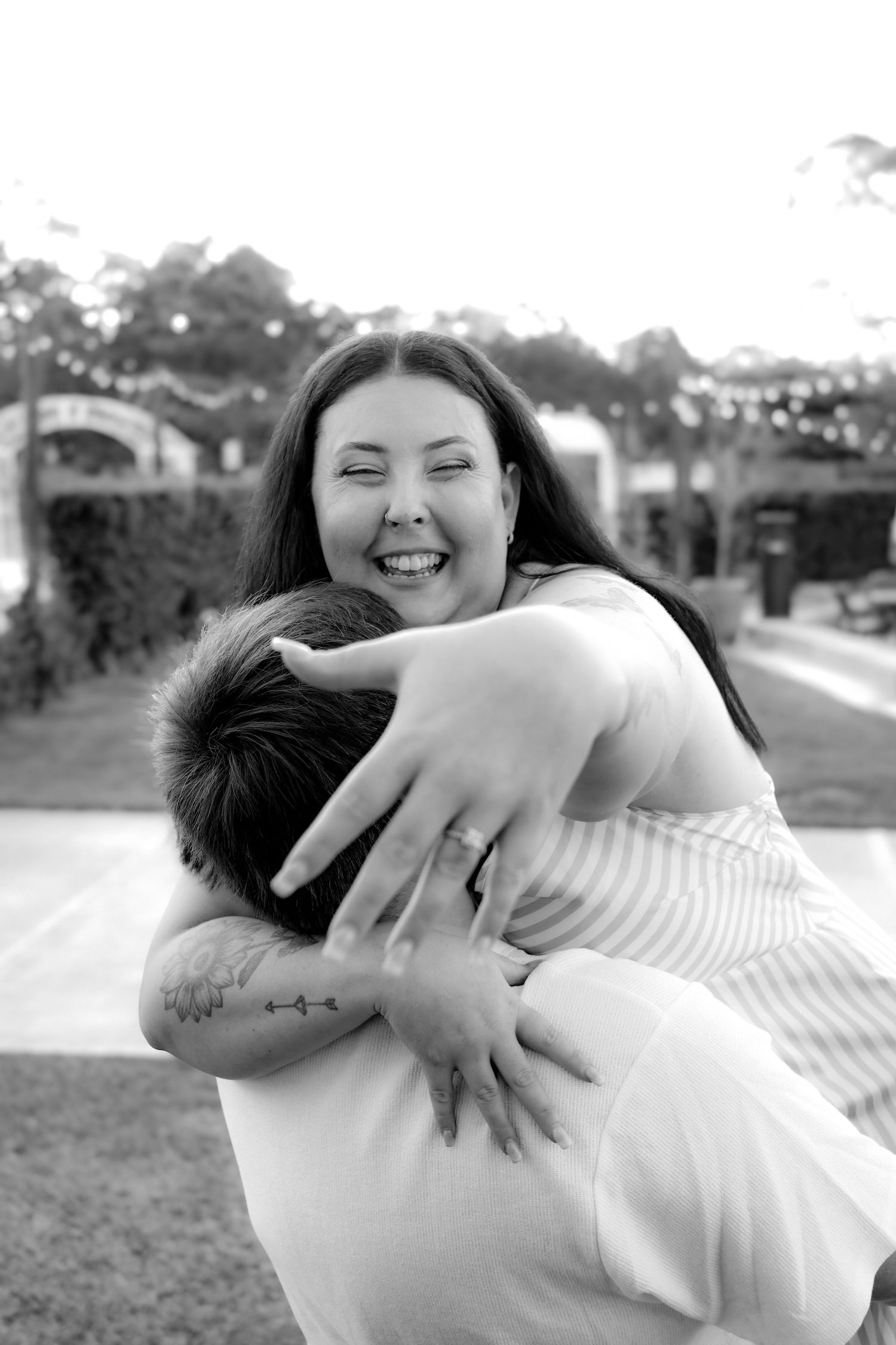 Black and white photo of a joyful woman hugging a man outdoors, laughing with eyes closed, showing engagement ring.