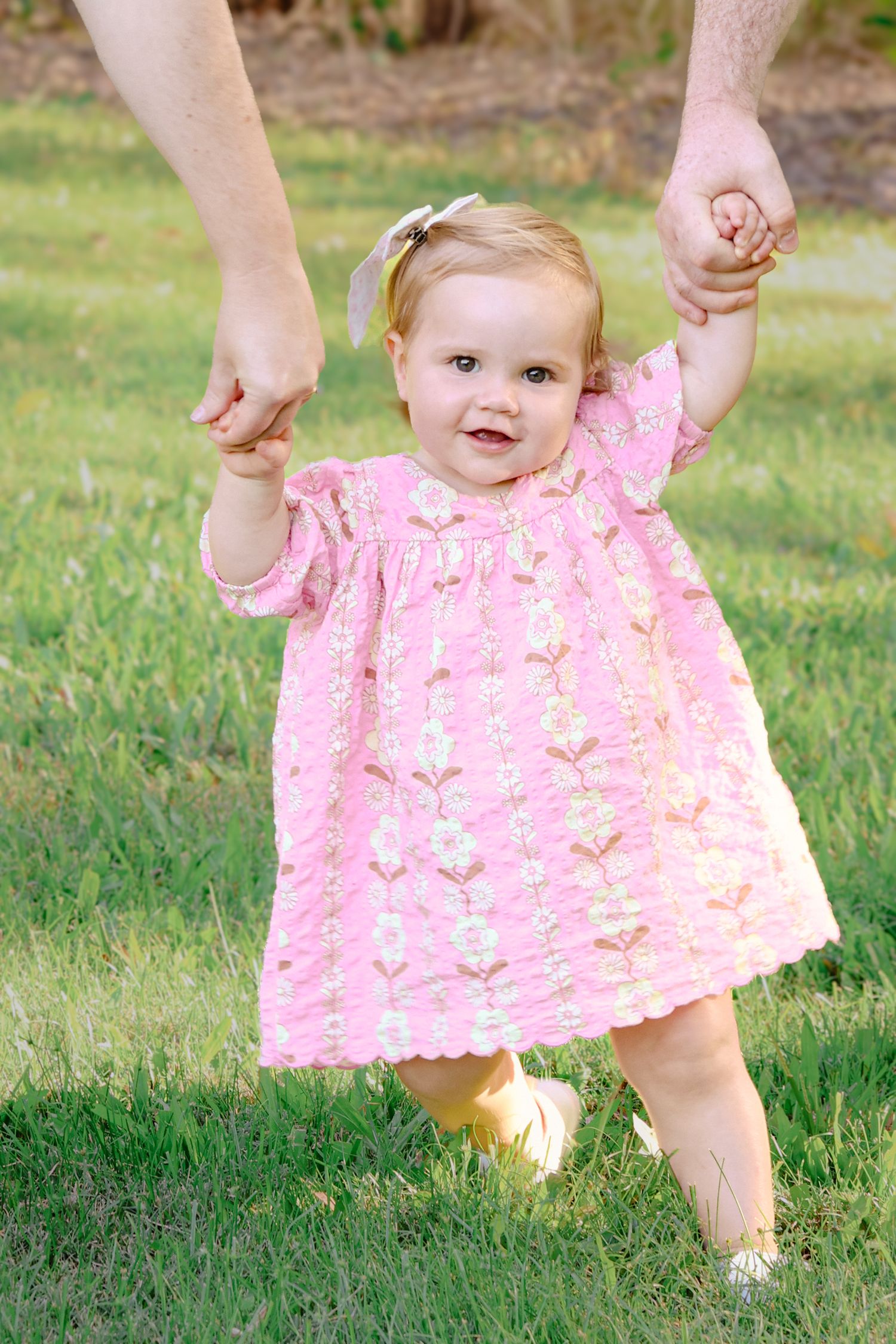 Adorable baby girl in pink floral dress learning to walk outdoors, held by adult hands on green grass.