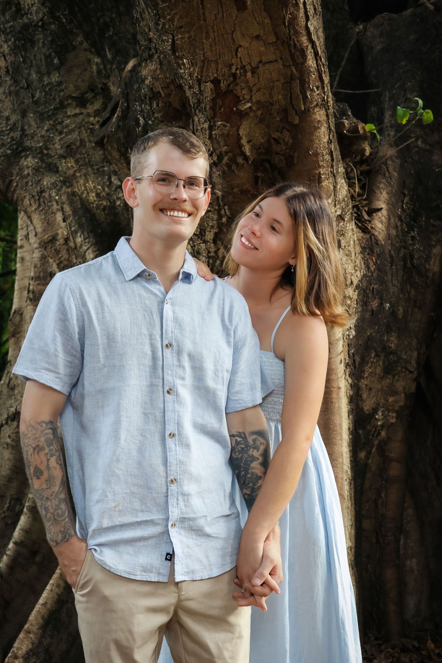 Smiling couple holding hands in front of a large tree, wearing matching light blue outfits with visible tattoos.