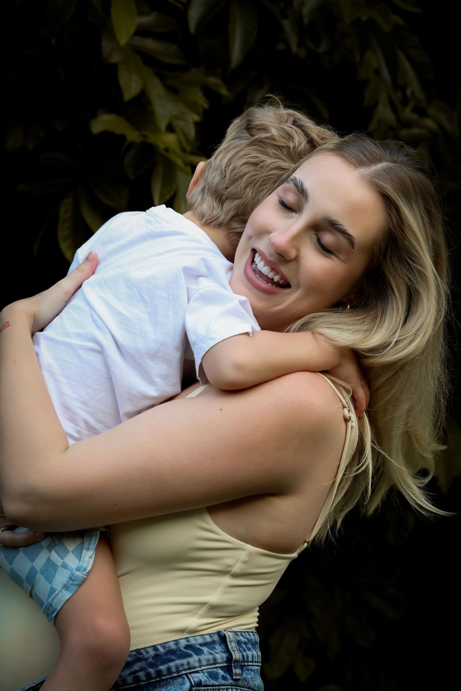 A smiling blonde woman joyfully hugs a young child outdoors, surrounded by lush green foliage.