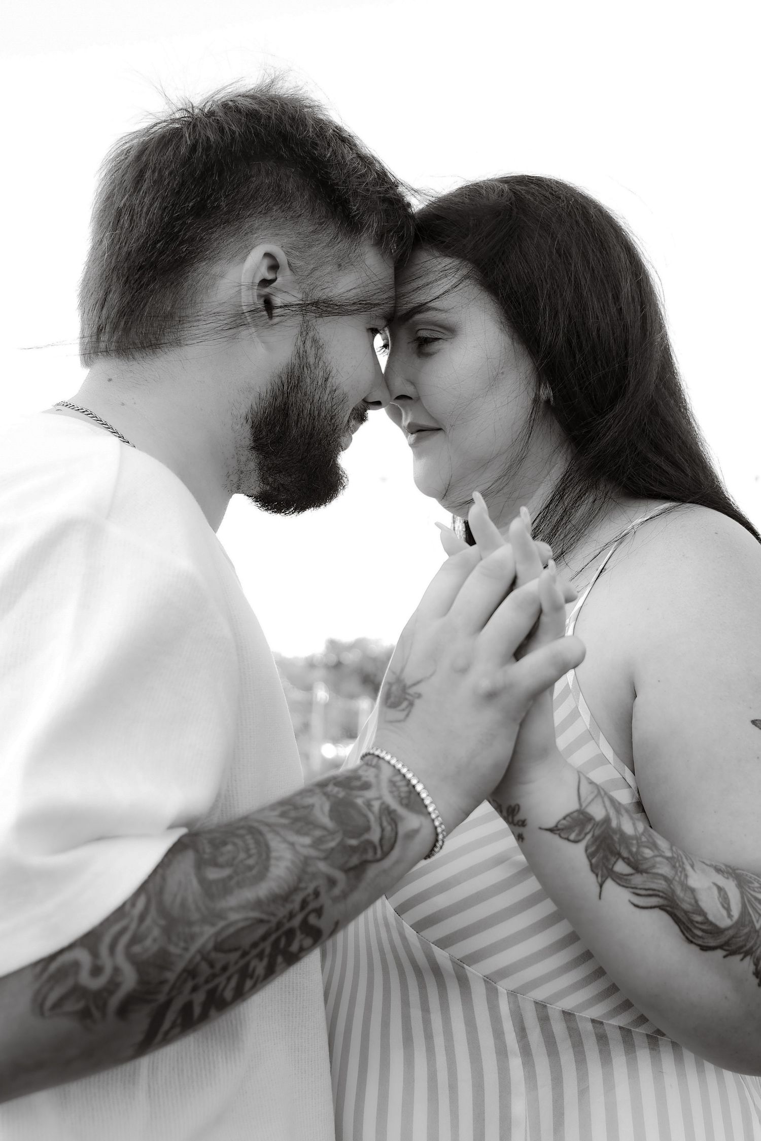 Black and white photo of tattooed couple touching foreheads romantically outdoors.
