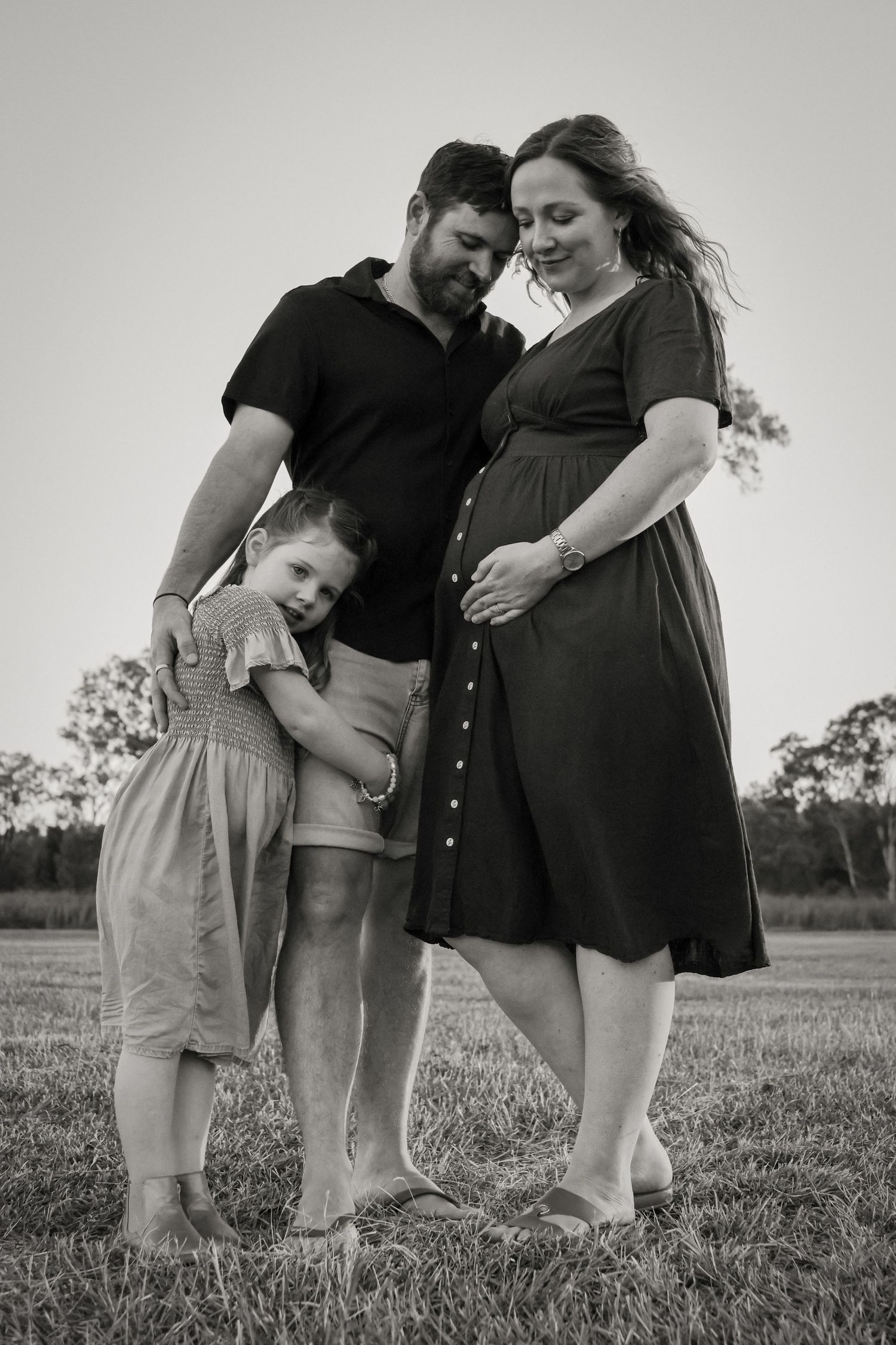 Black and white maternity photo of a pregnant woman, man, and young girl standing together in a field.