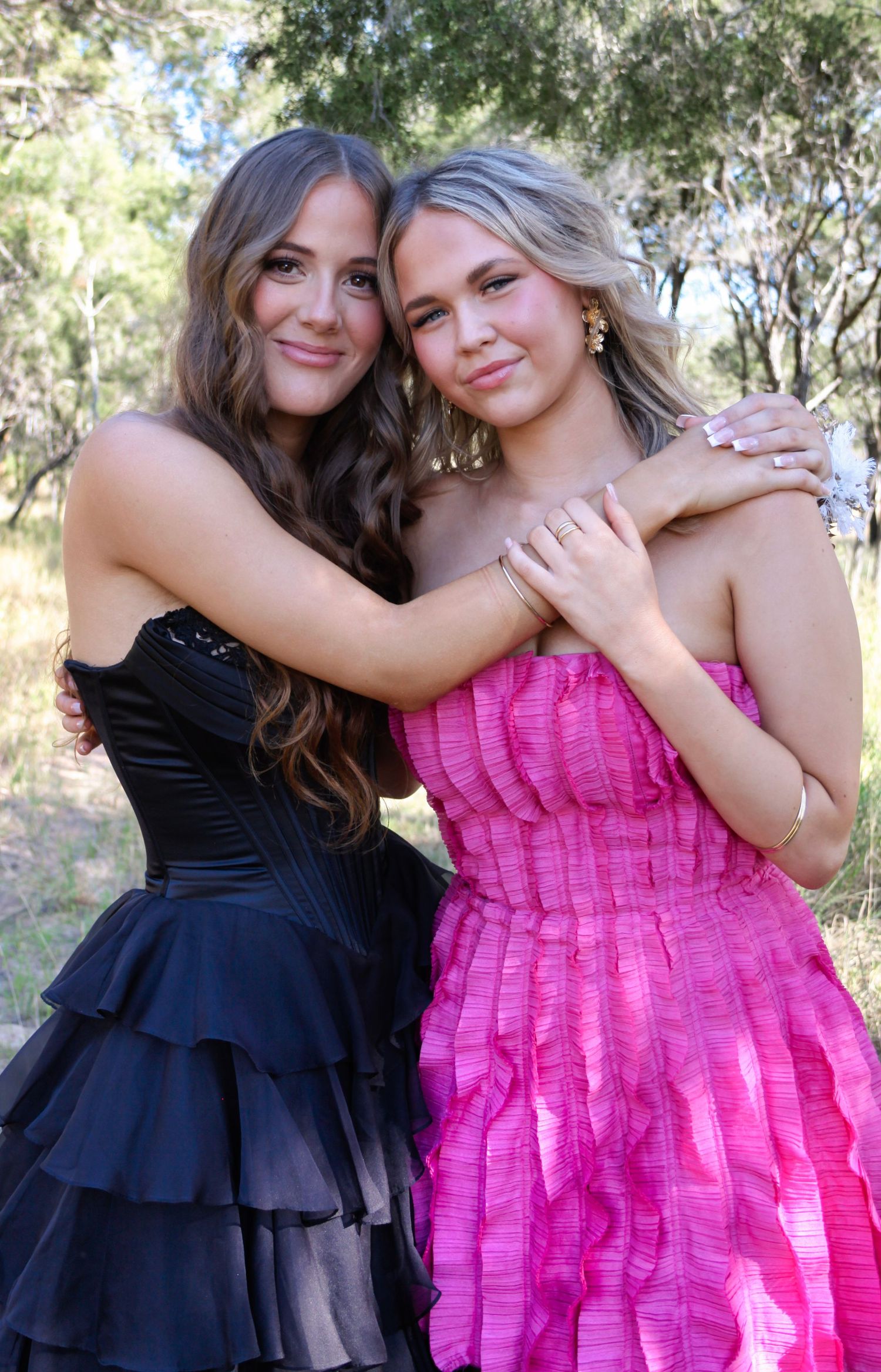 Two friends pose together outdoors wearing contrasting black and pink formal dresses in a natural setting.