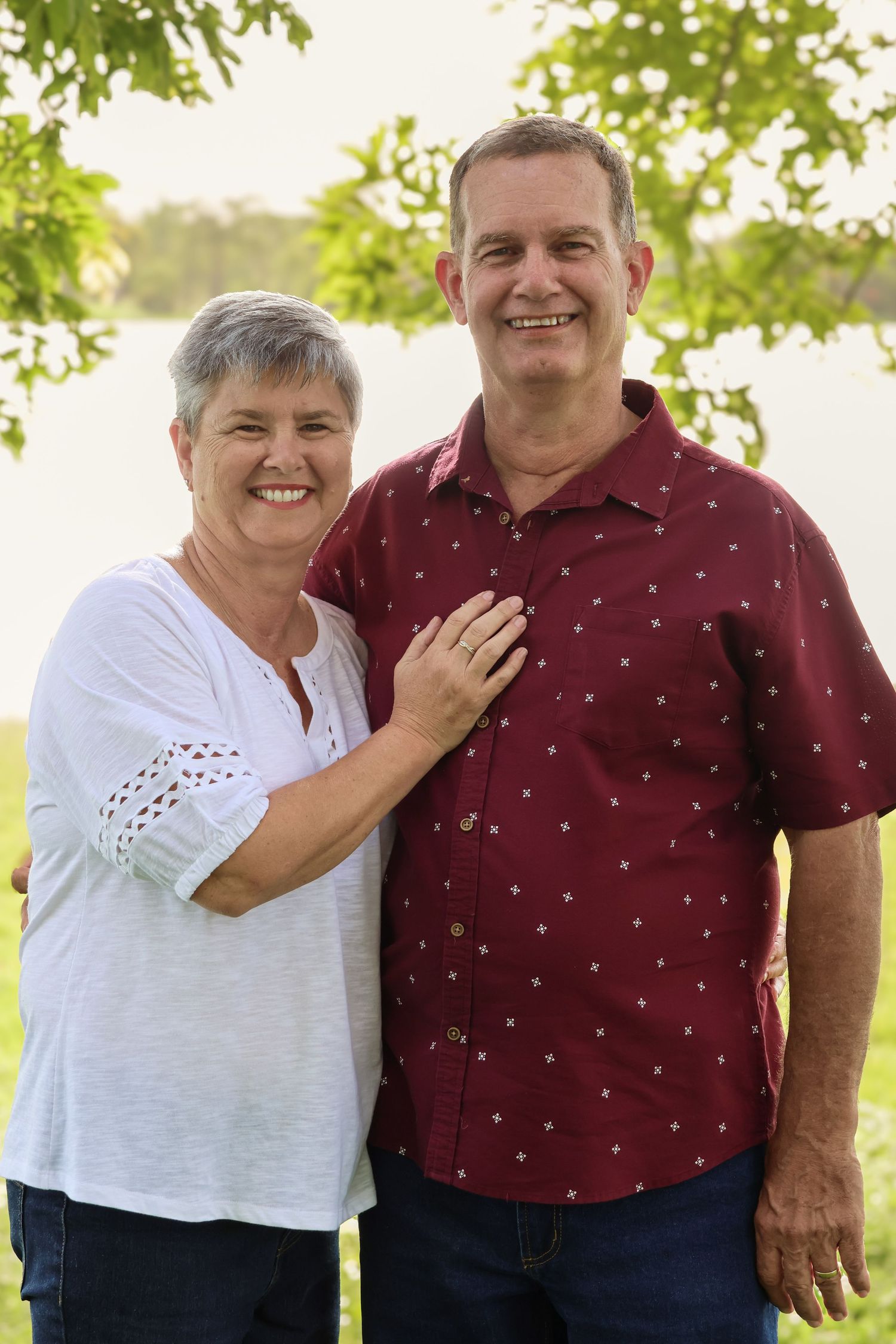 A smiling middle-aged couple poses outdoors near a lake, woman in white top and man in maroon shirt.