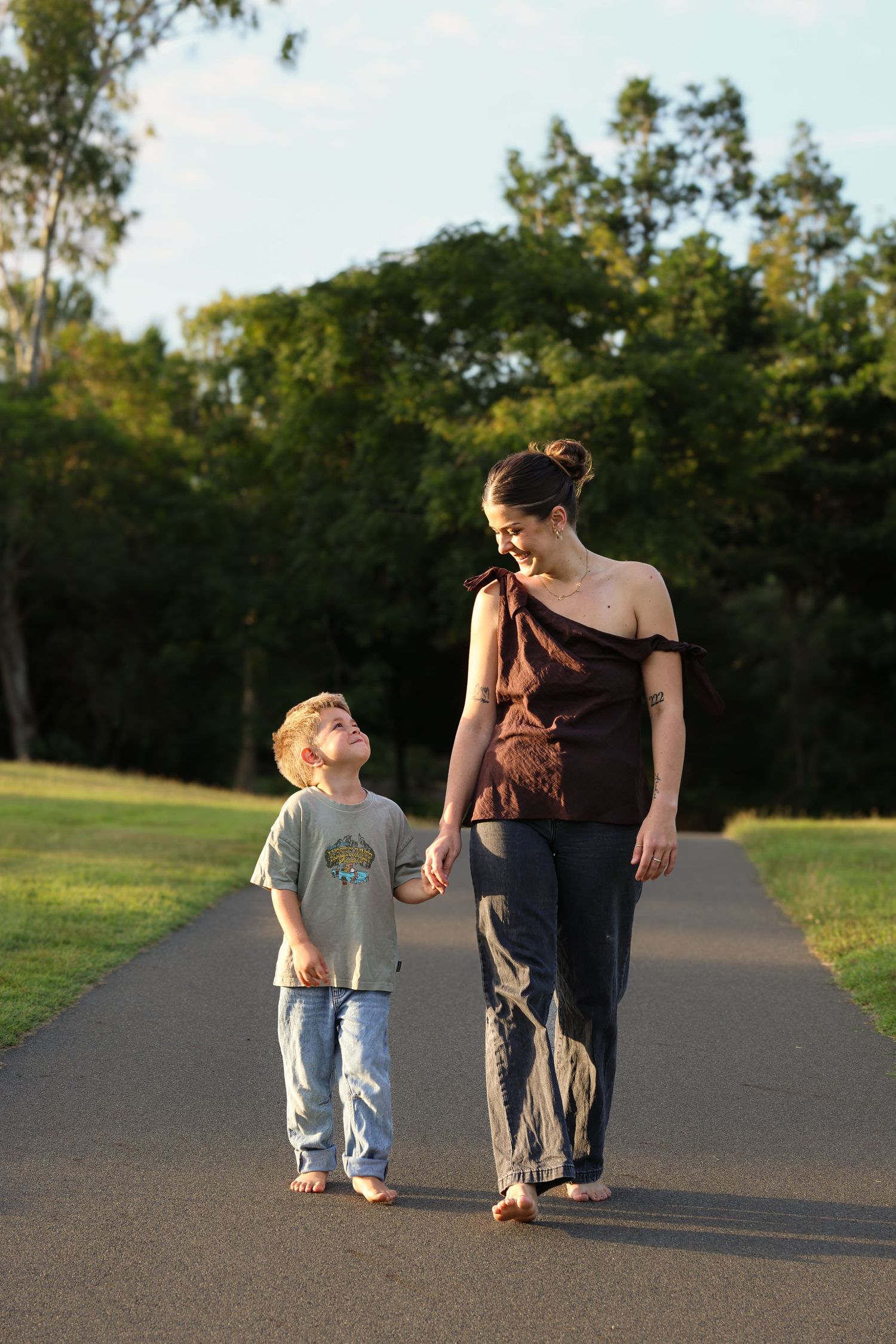 A mother and young son walk hand-in-hand along a park path, smiling at each other on a sunny day.