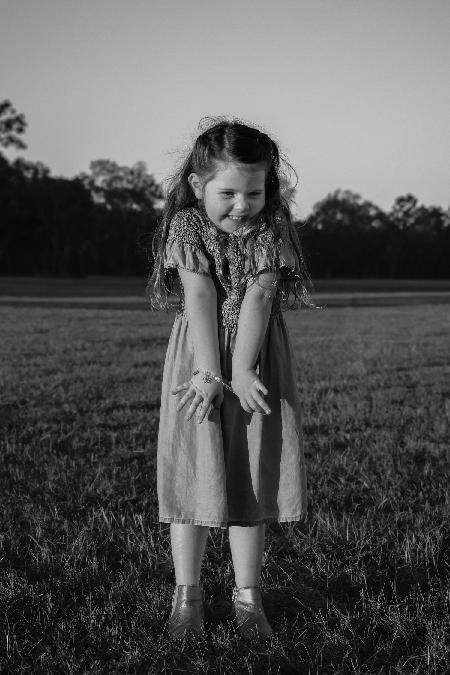 Black and white photo of a young girl in a dress smiling and standing in an open grassy field at dusk.