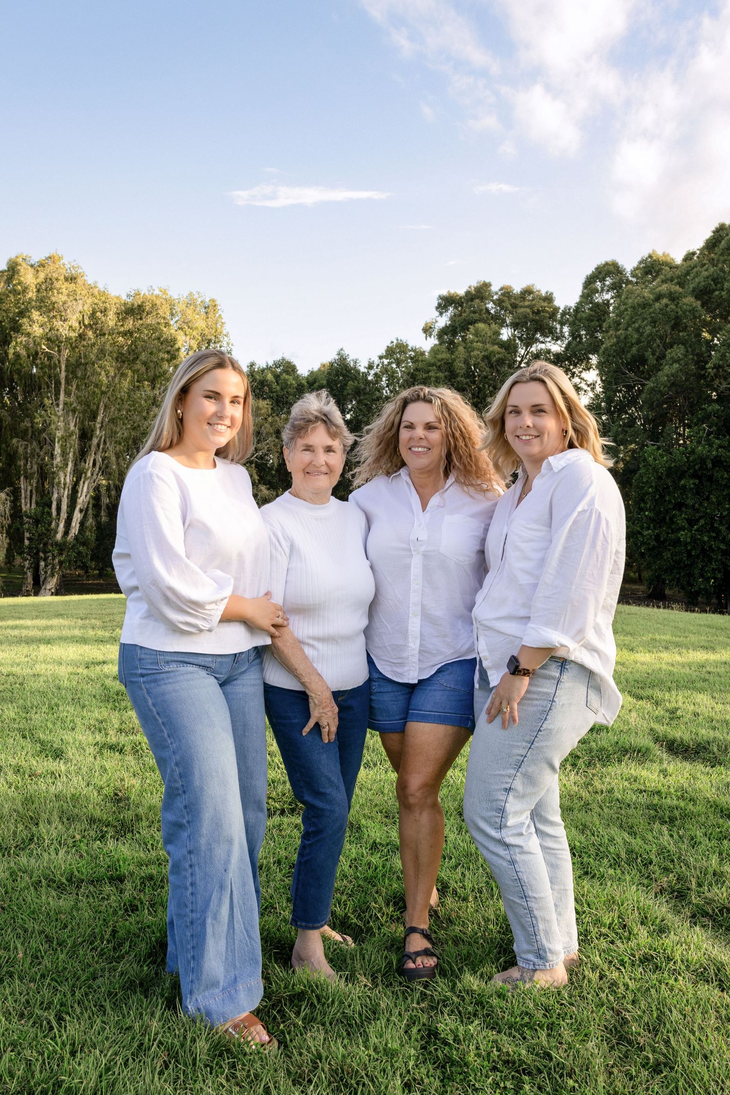 Four women in white tops and jeans pose together on green grass with trees and blue sky behind them.