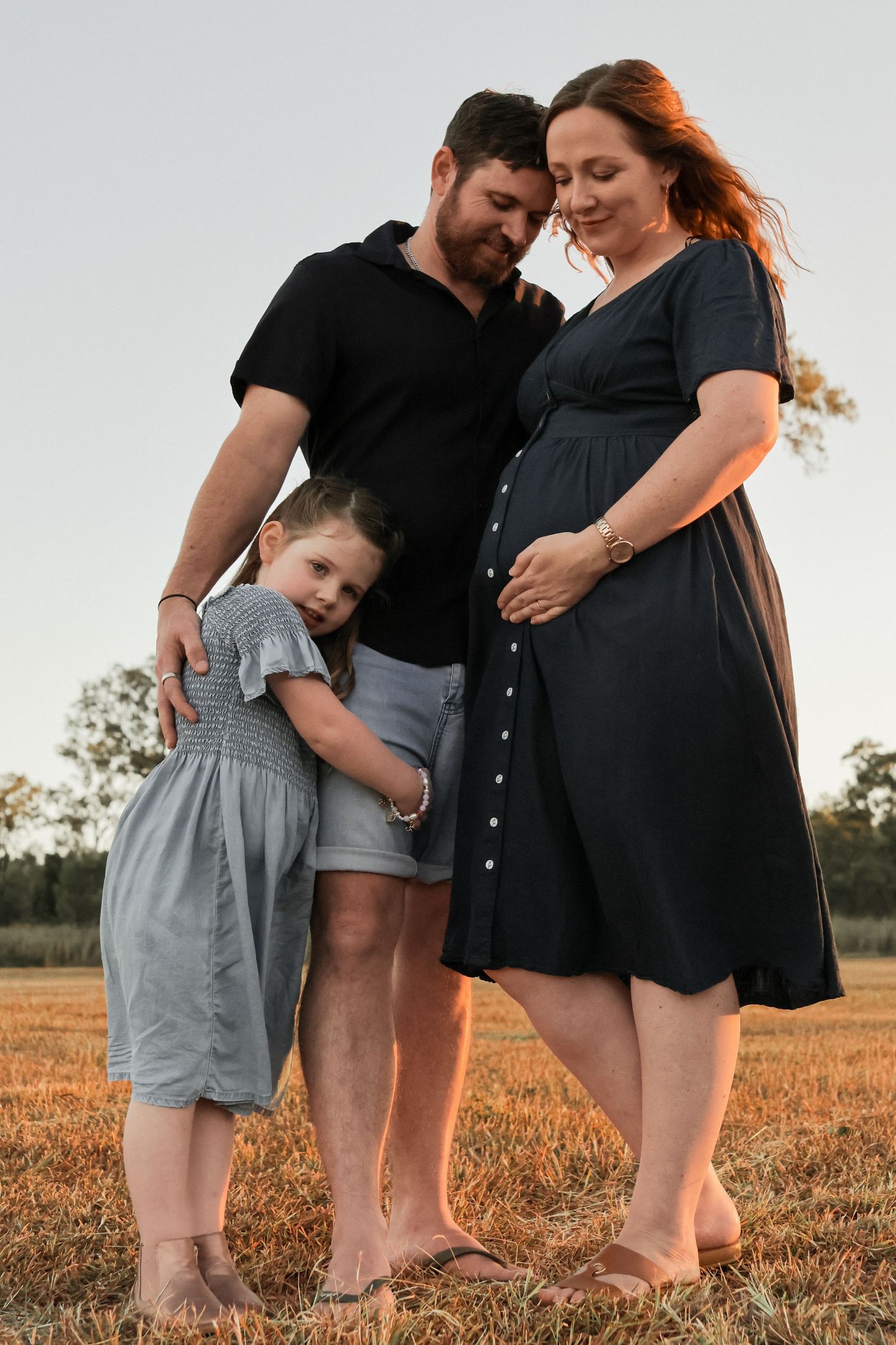 Pregnant woman in navy dress with partner and young daughter posing in golden field at sunset.