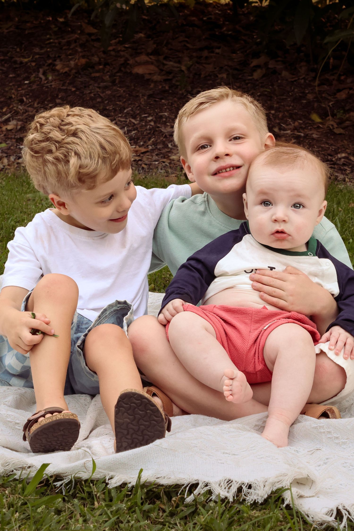 Three young blonde siblings sit together on a blanket outdoors, two boys hugging a smiling baby in red shorts.