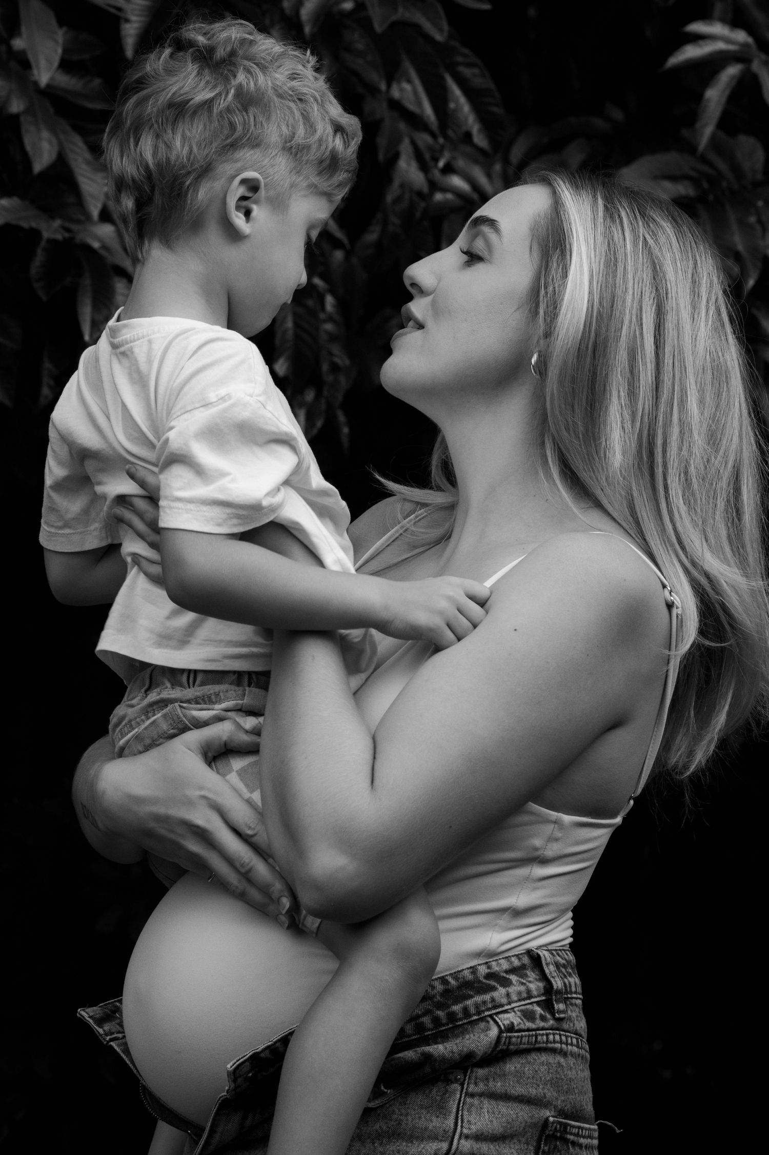 Black and white photo of a smiling blonde pregnant woman holding a young toddler boy outdoors.