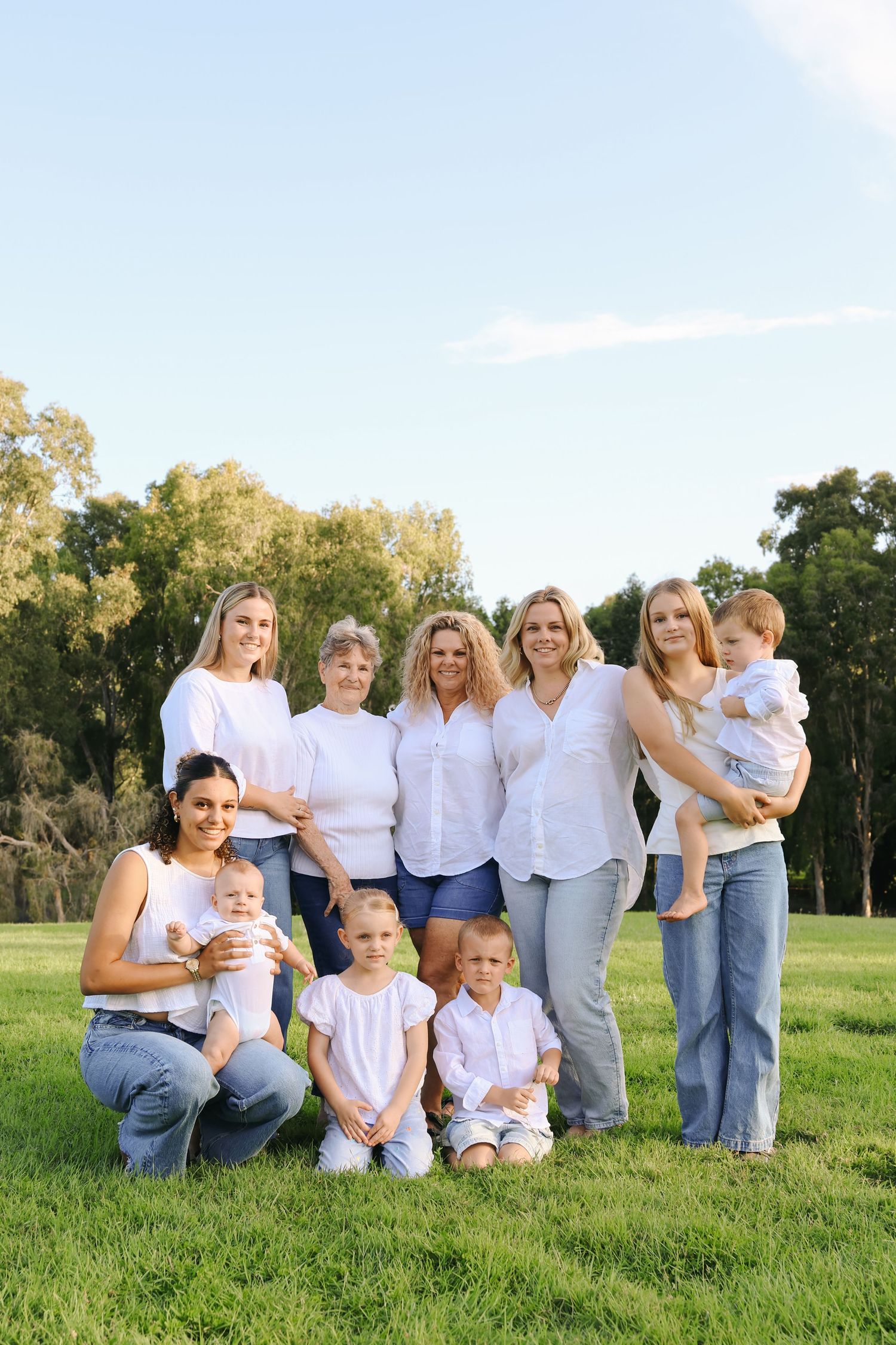 Multi-generational family group in white tops and jeans posing outdoors on green grass with trees and blue sky.