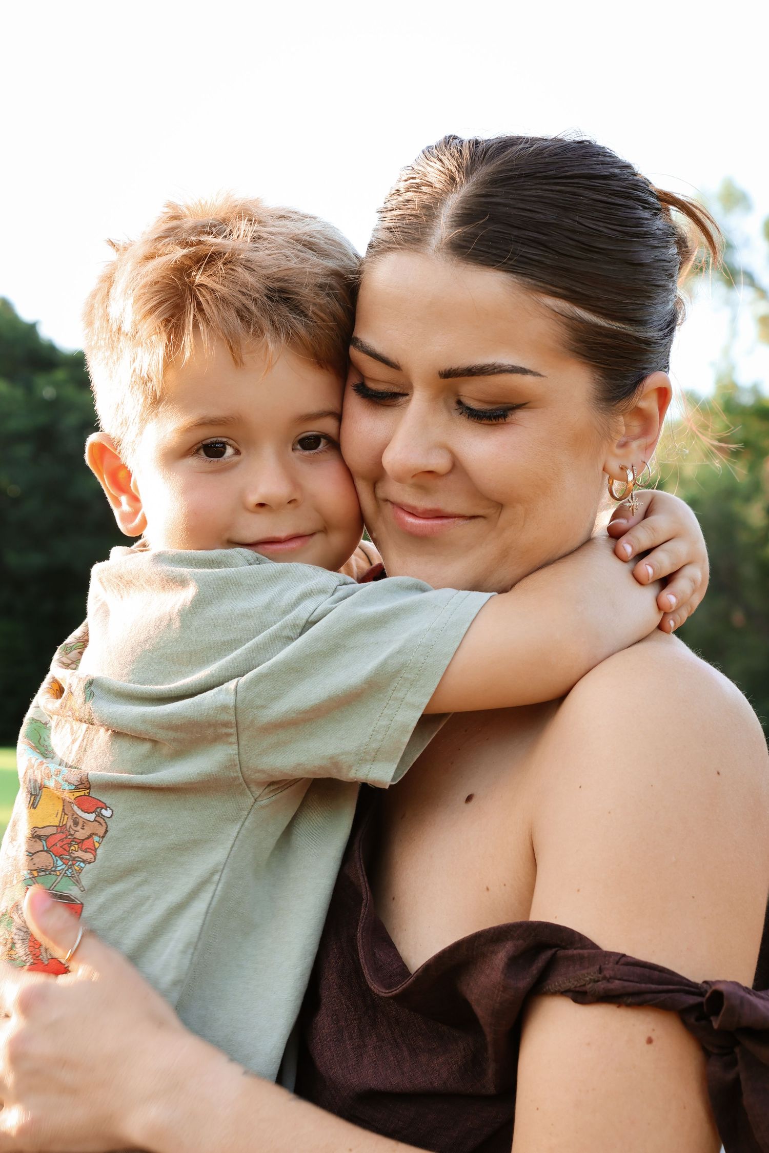 A young boy hugs a smiling dark-haired woman outdoors, both sharing a warm embrace in natural light.