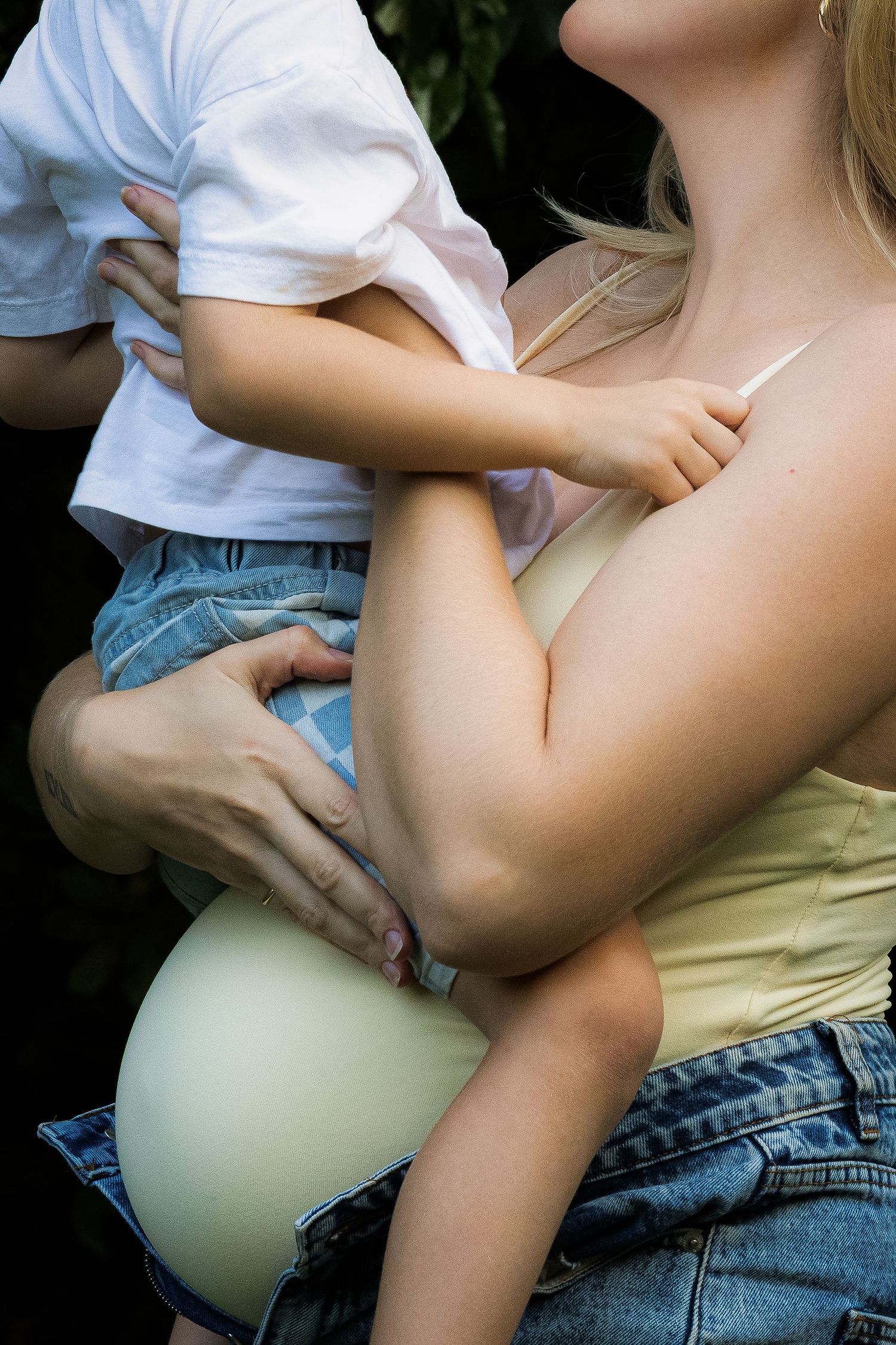 Pregnant woman holding a young child outdoors, wearing a yellow top and jeans, cradling her baby bump.