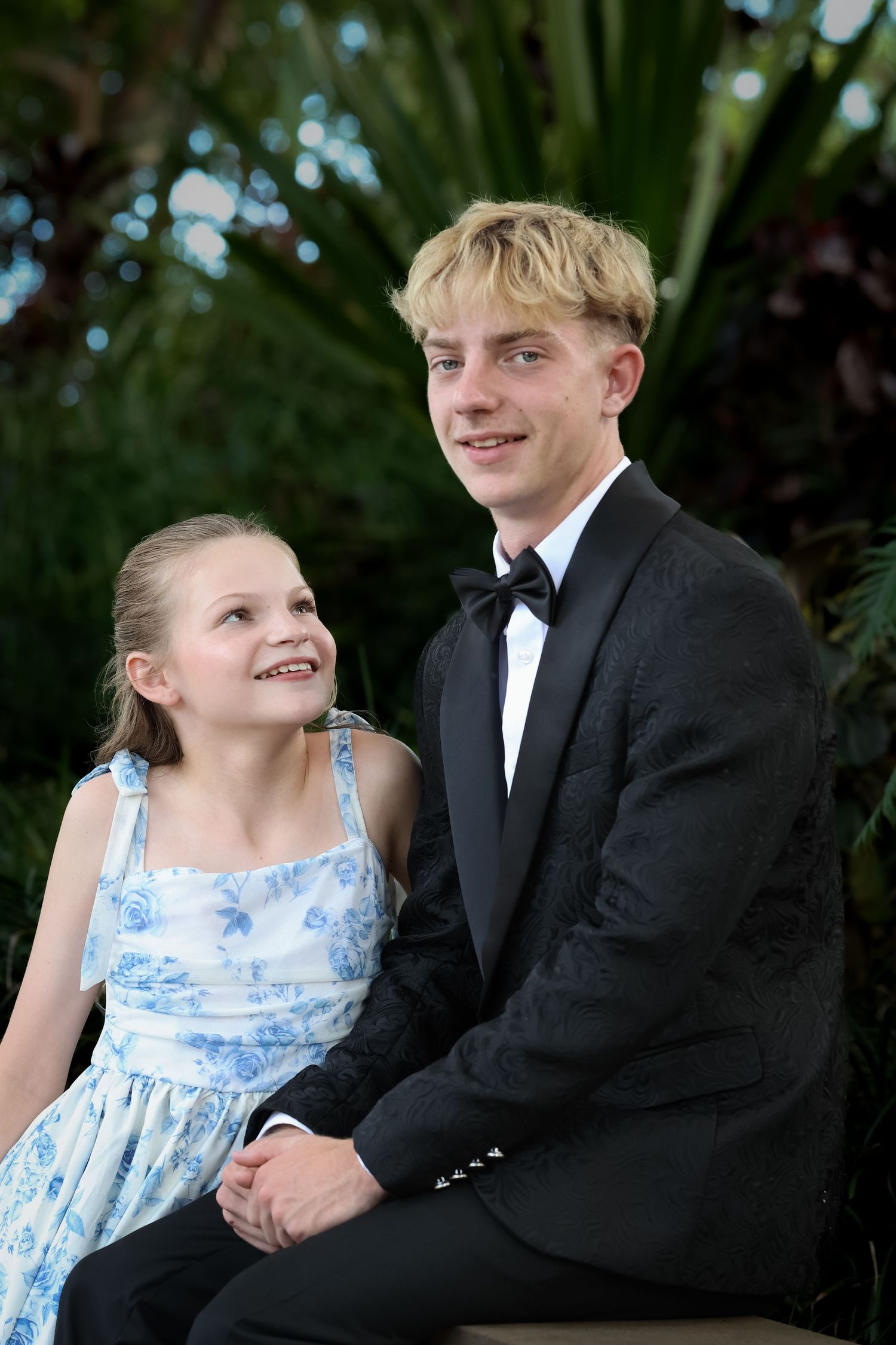 A teenage boy in a black tuxedo sits with a young girl in a blue floral dress, smiling outdoors.