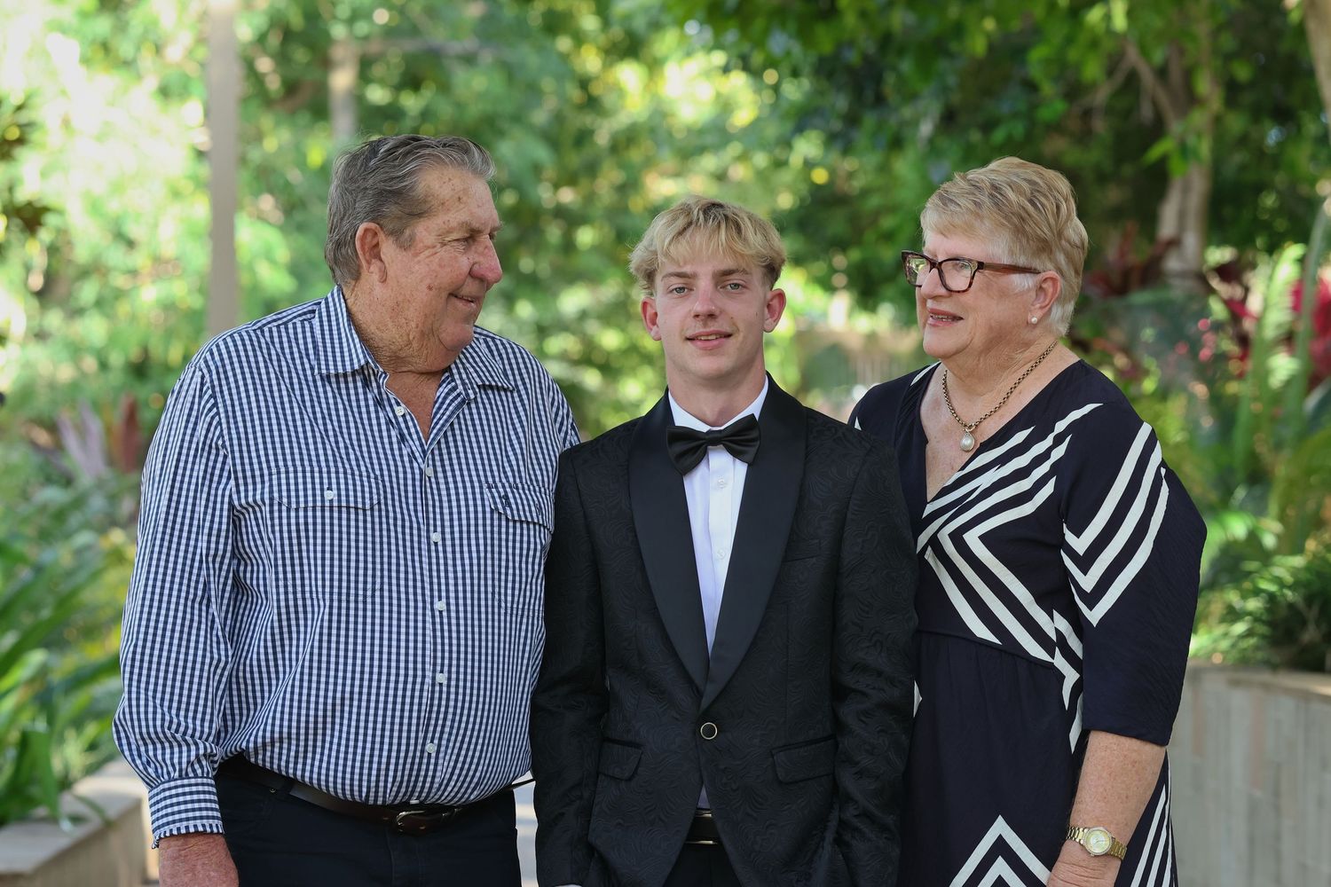 A teenage boy in a tuxedo poses with two smiling adults outdoors among lush green trees.