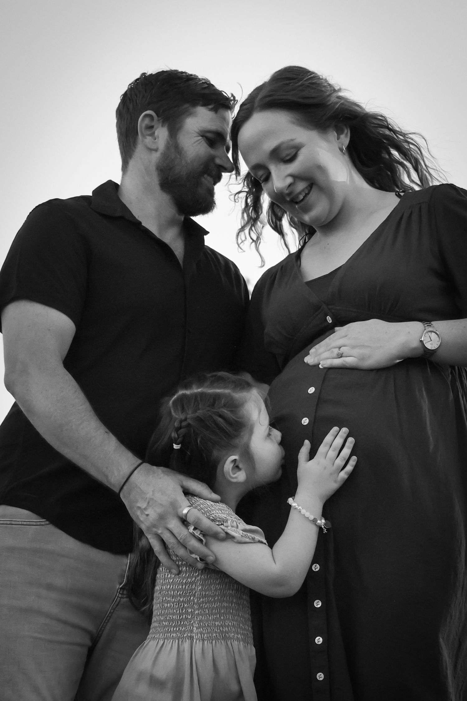 Black and white maternity photo of pregnant woman, man, and young girl touching baby bump outdoors.