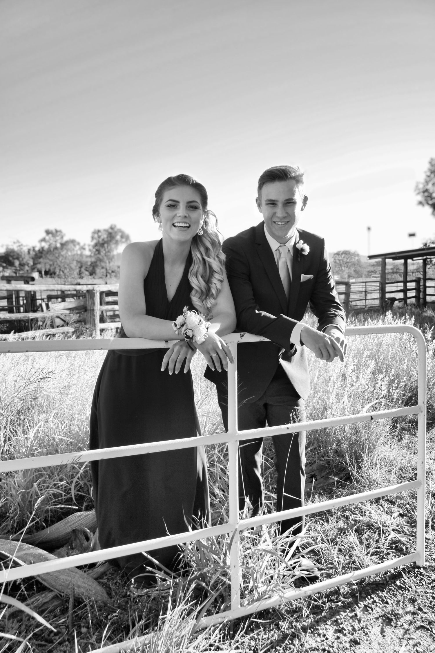 Black and white photo of couple leaning on wooden fence in countryside setting.