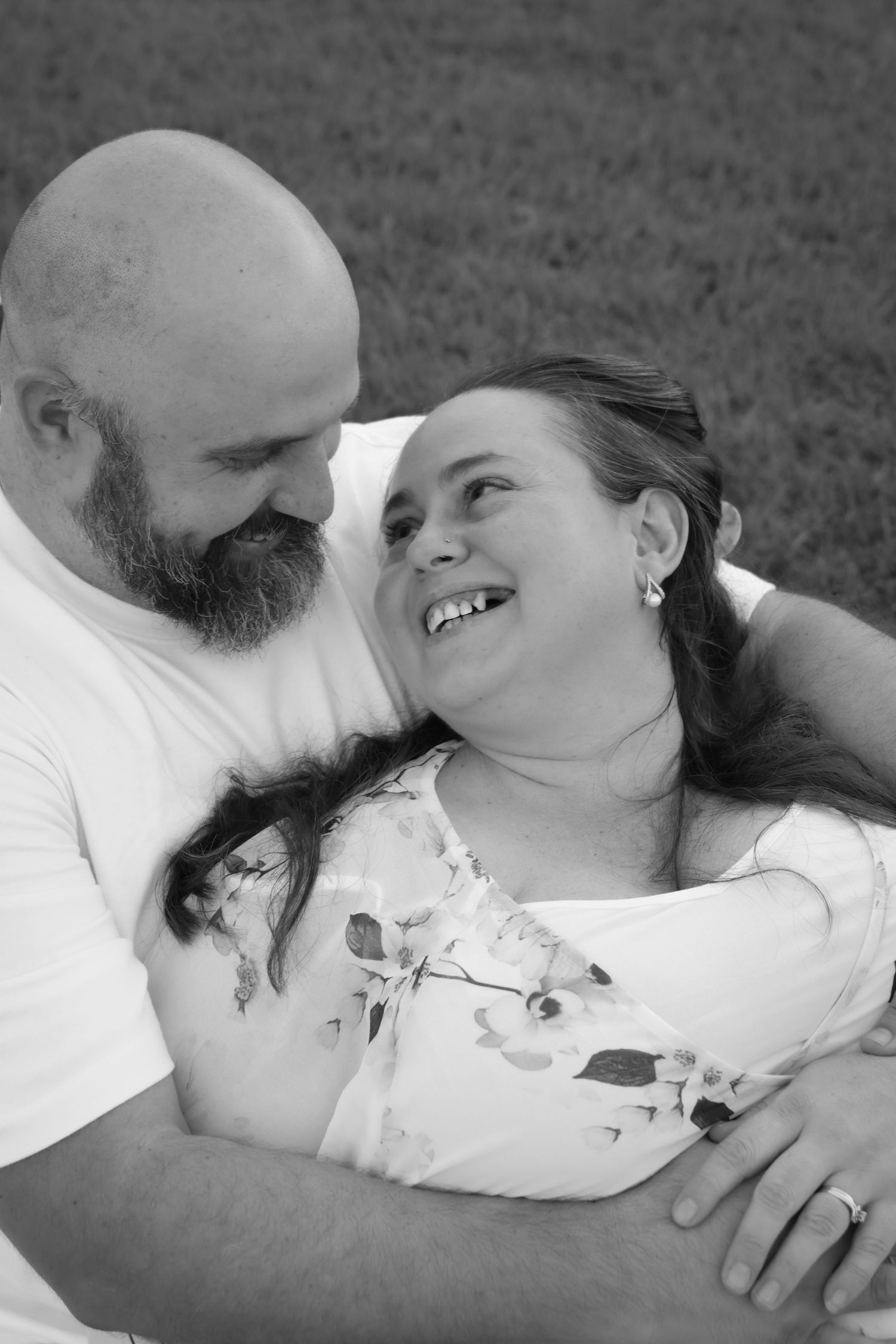 Black and white photo of a smiling couple embracing outdoors, bald bearded man hugging a laughing woman in floral dress.
