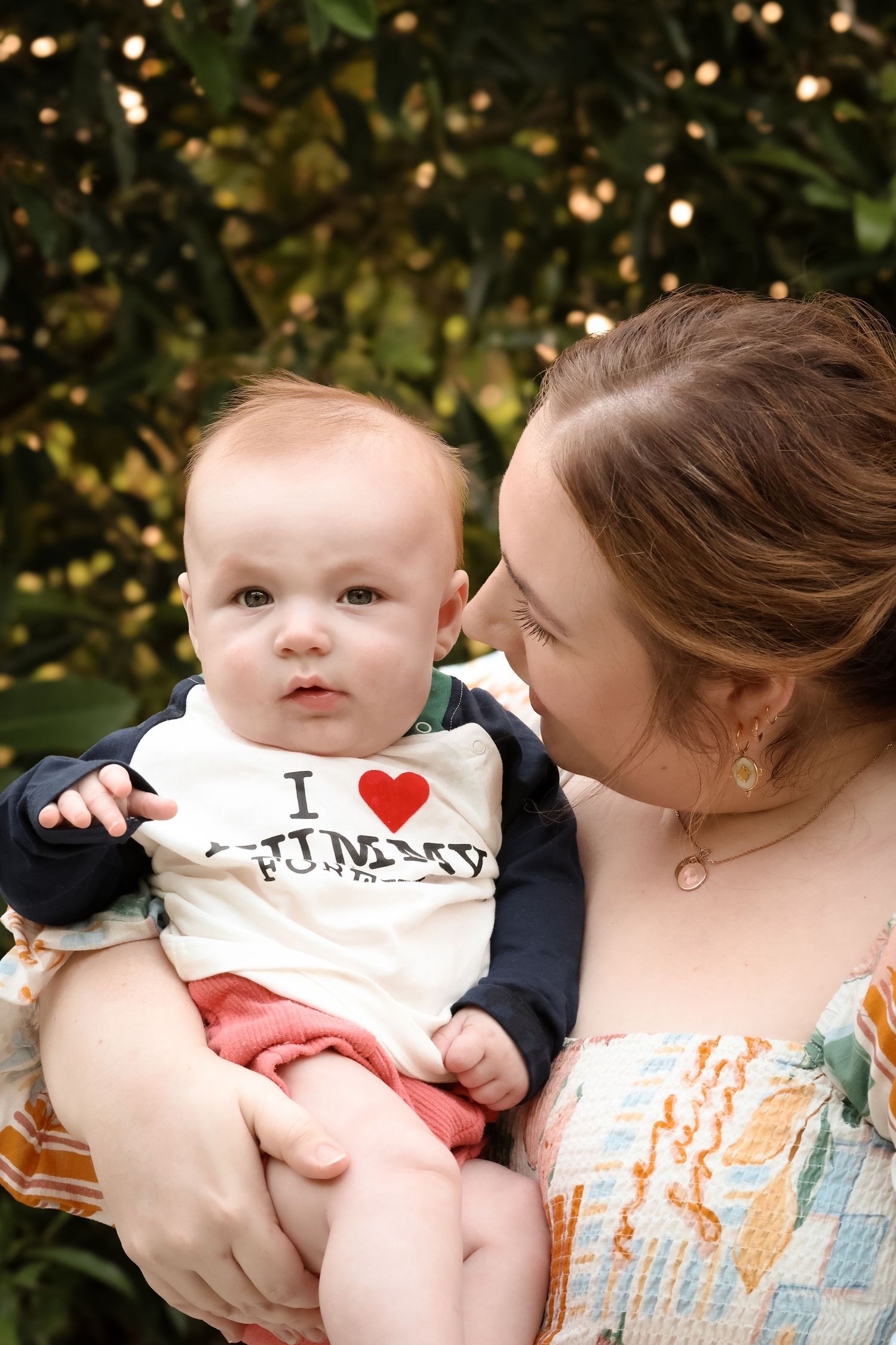 A smiling mother holds her baby wearing an I Love Mommy shirt outdoors with greenery in the background.