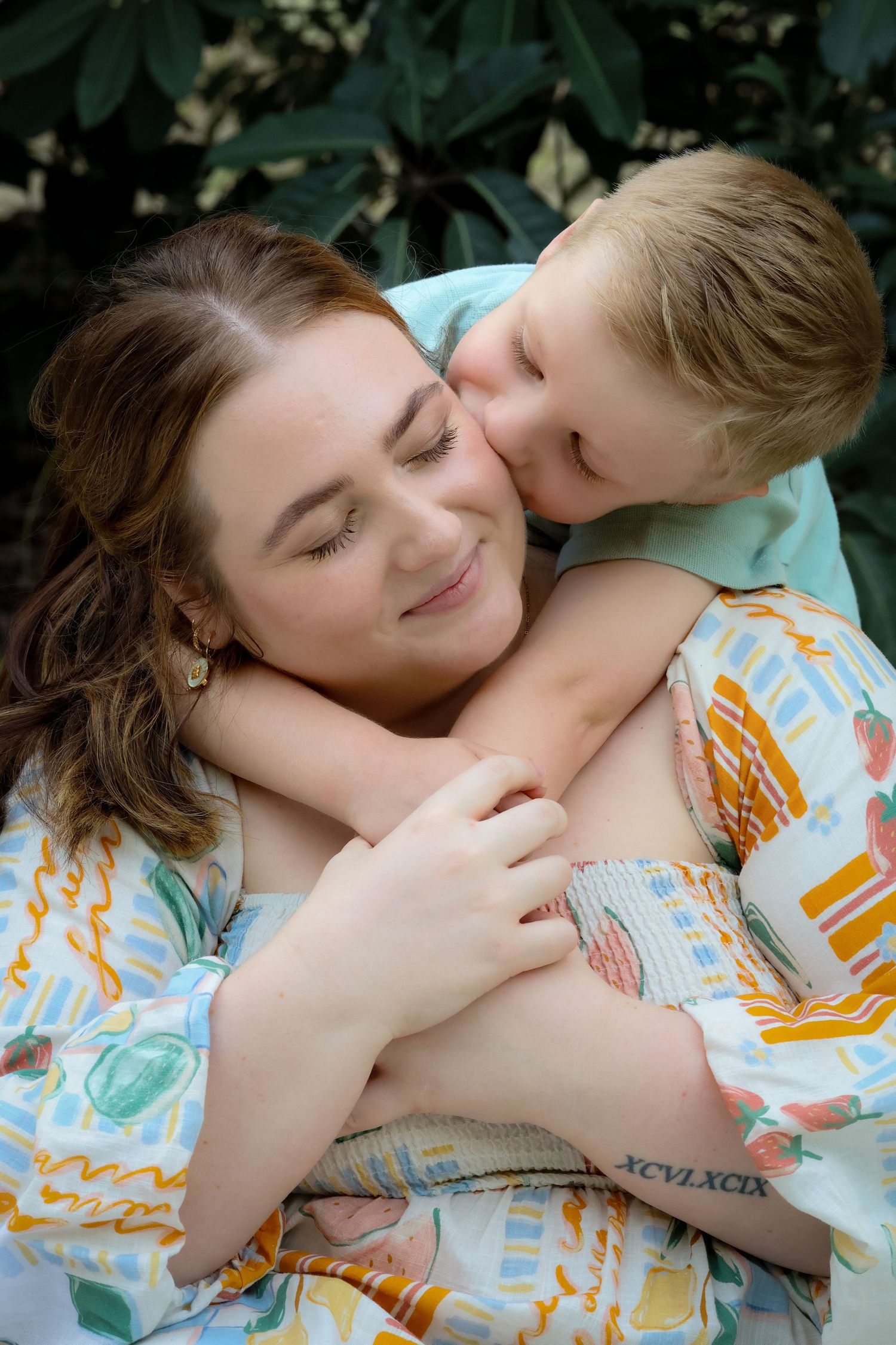 A young boy hugs and kisses a smiling woman outdoors, both wearing colorful patterned clothing near green foliage.