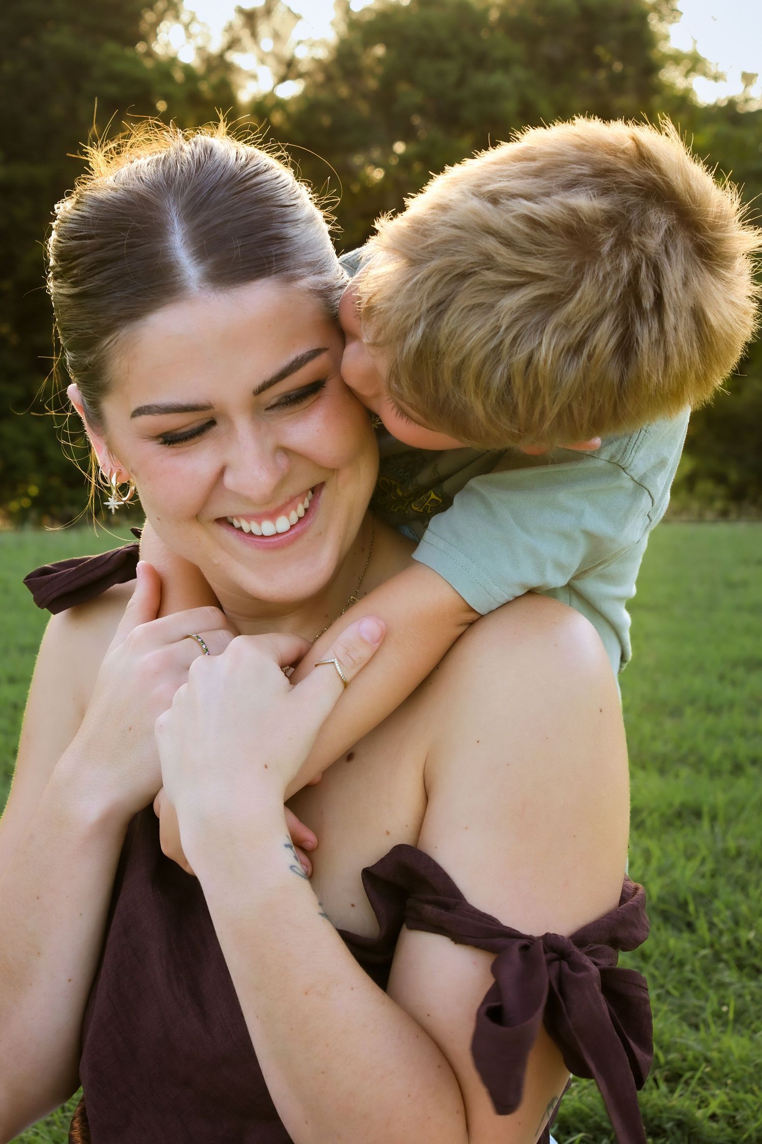 A smiling woman enjoys a sweet kiss from a young boy hugging her from behind outdoors.