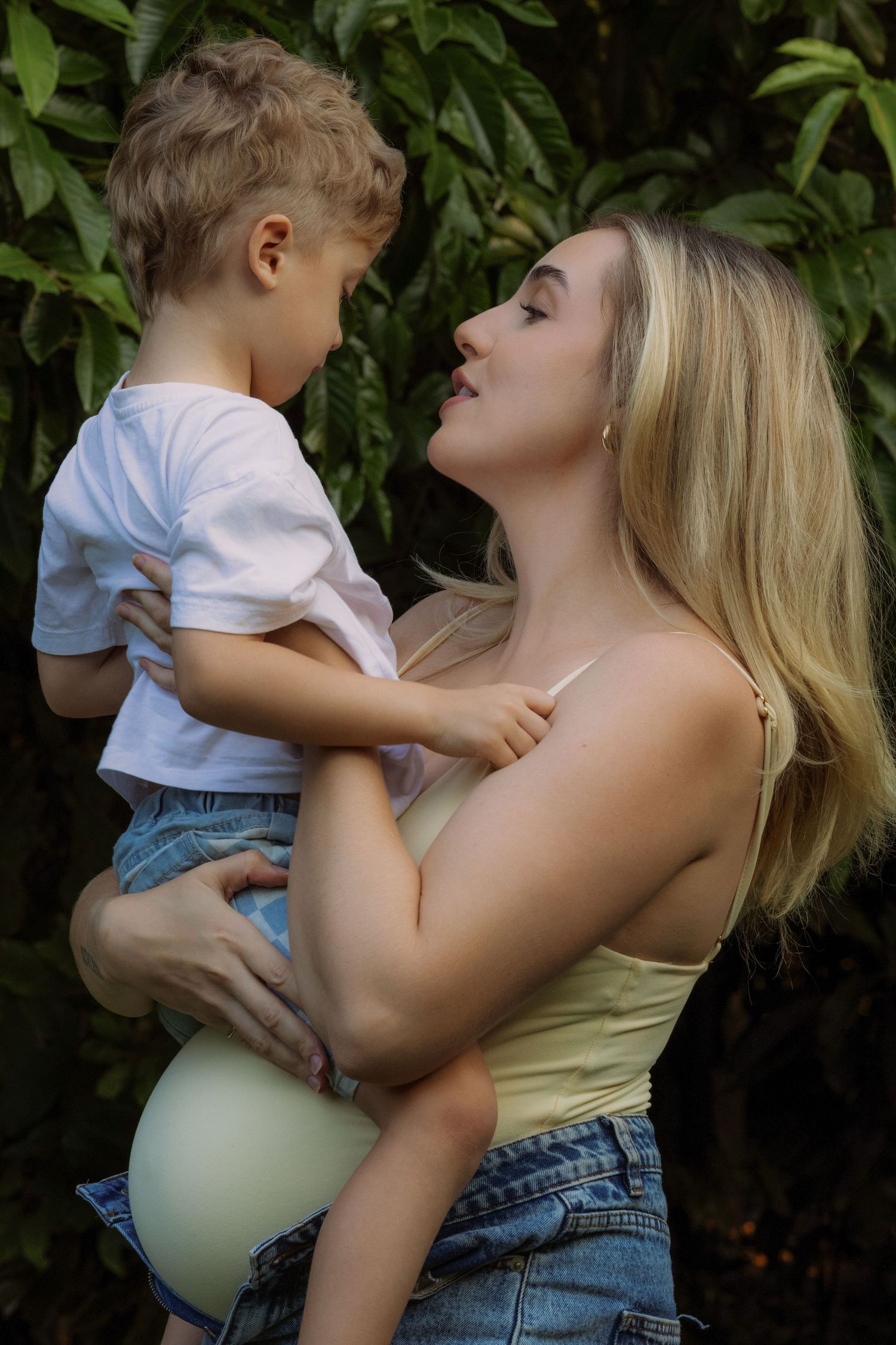 Pregnant blonde woman holding a young boy outdoors, surrounded by lush green tropical foliage.