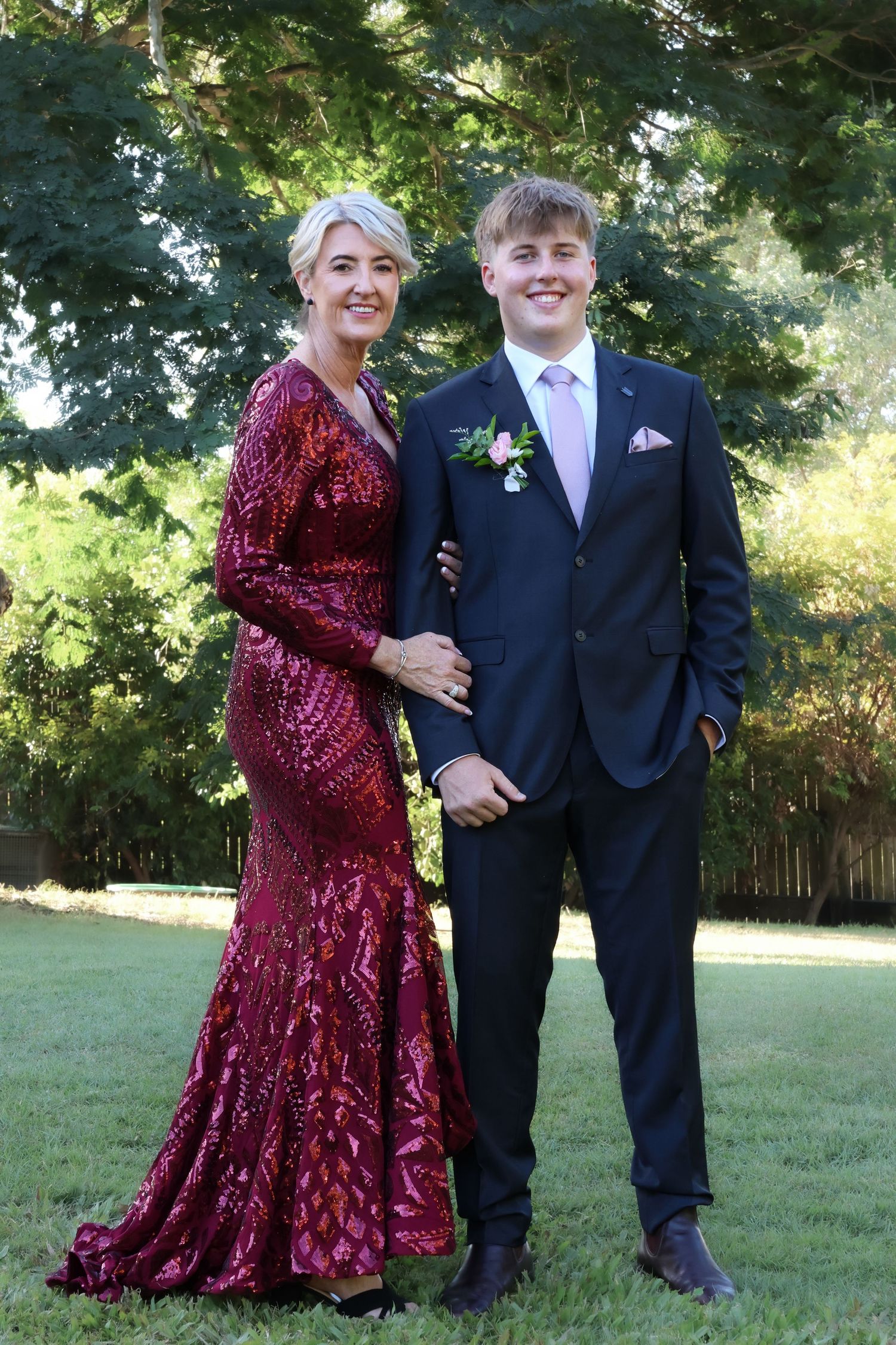 A young man in a navy suit with boutonniere poses with a woman in a red sequin gown outdoors on green grass.