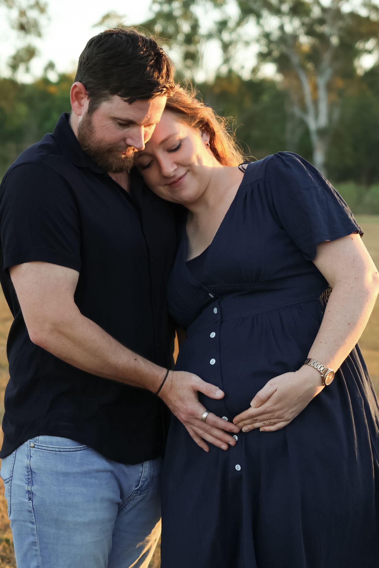 Couple sharing a tender moment outdoors, man embracing pregnant woman in navy dress, golden hour light.