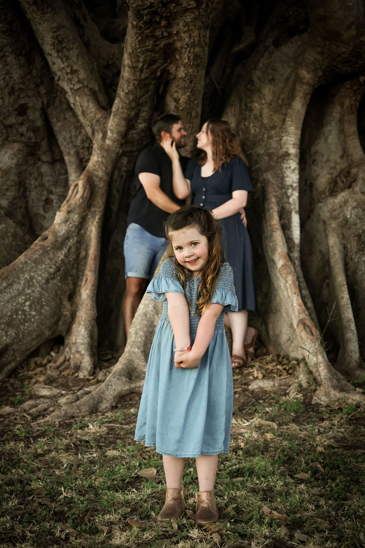 Young girl in blue dress smiles at camera while couple shares romantic moment by large tree roots behind her.