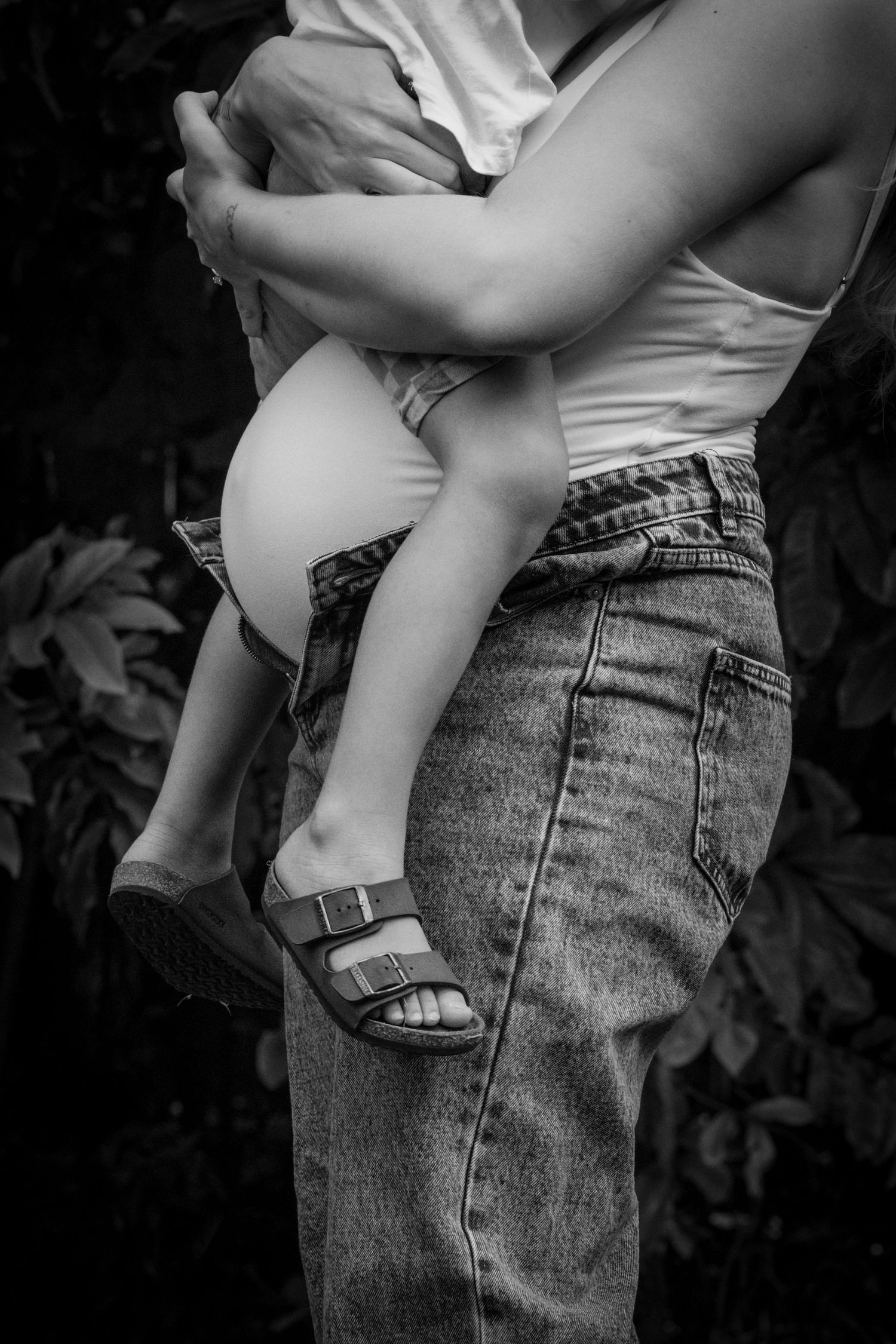 Black and white photo of a mother holding a young child outdoors, wearing jeans and Birkenstock sandals.
