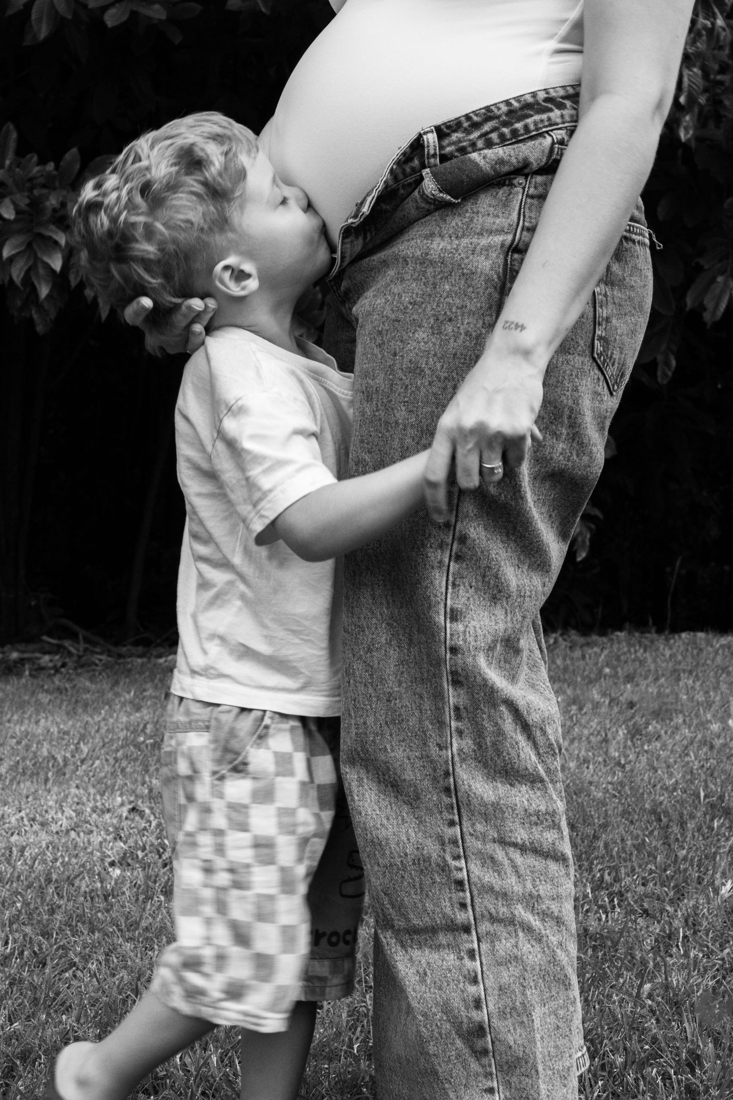 Black and white photo of a young boy kissing his pregnant mother's belly outdoors.