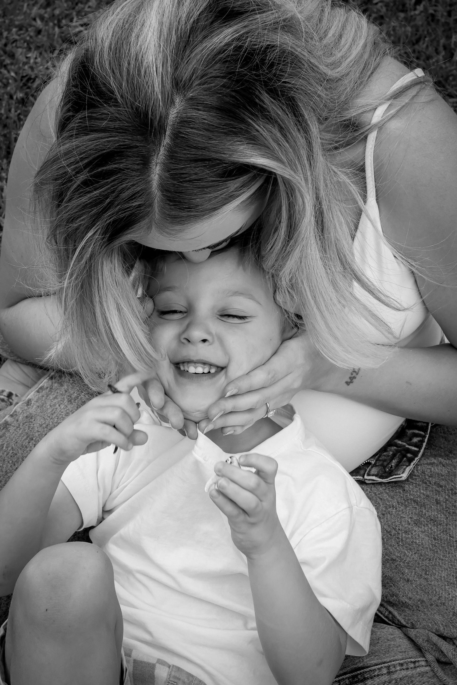 Black and white photo of a laughing child being hugged and kissed by a smiling woman outdoors.