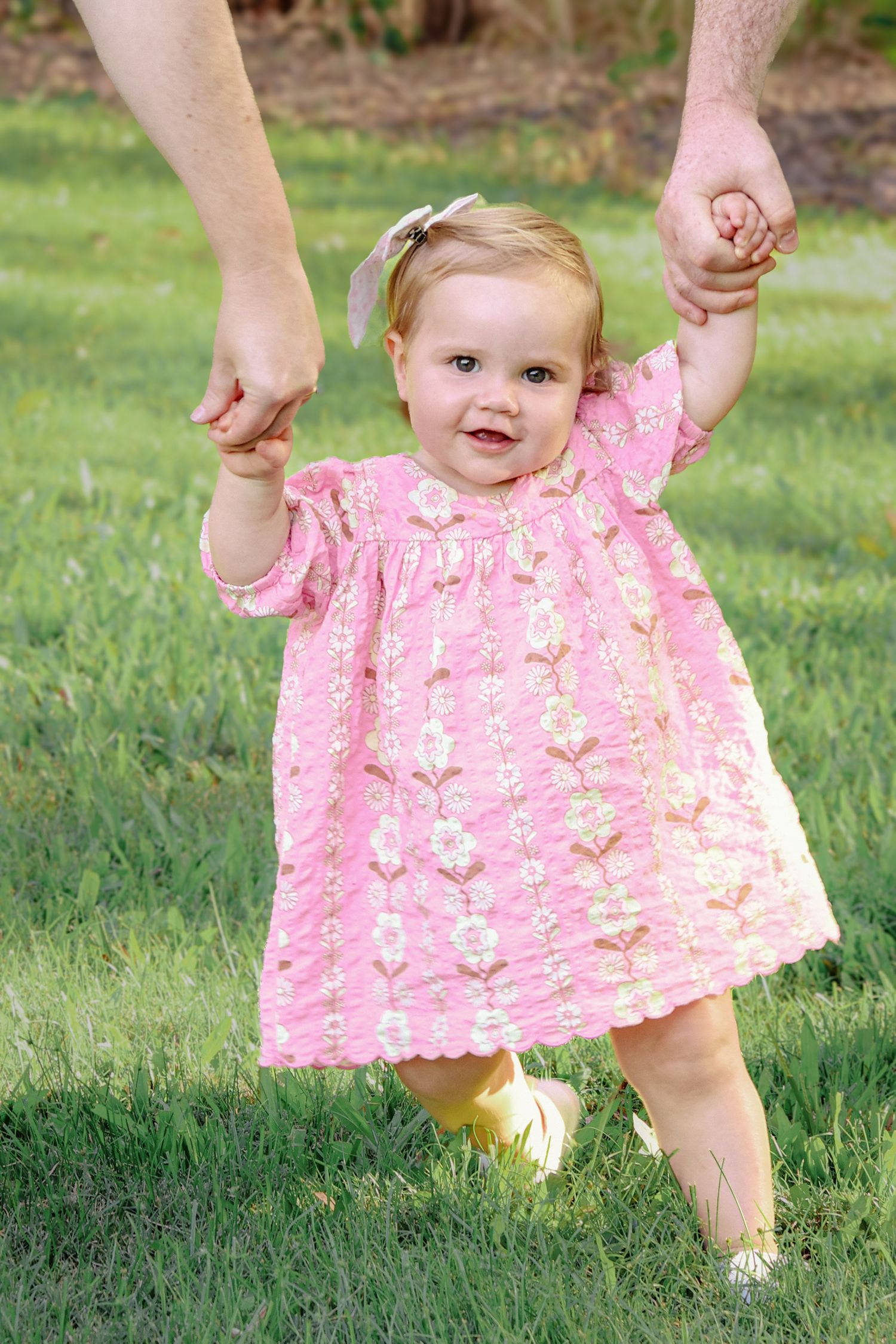 Toddler girl in pink floral dress learning to walk outdoors, held by adult hands on green grass.