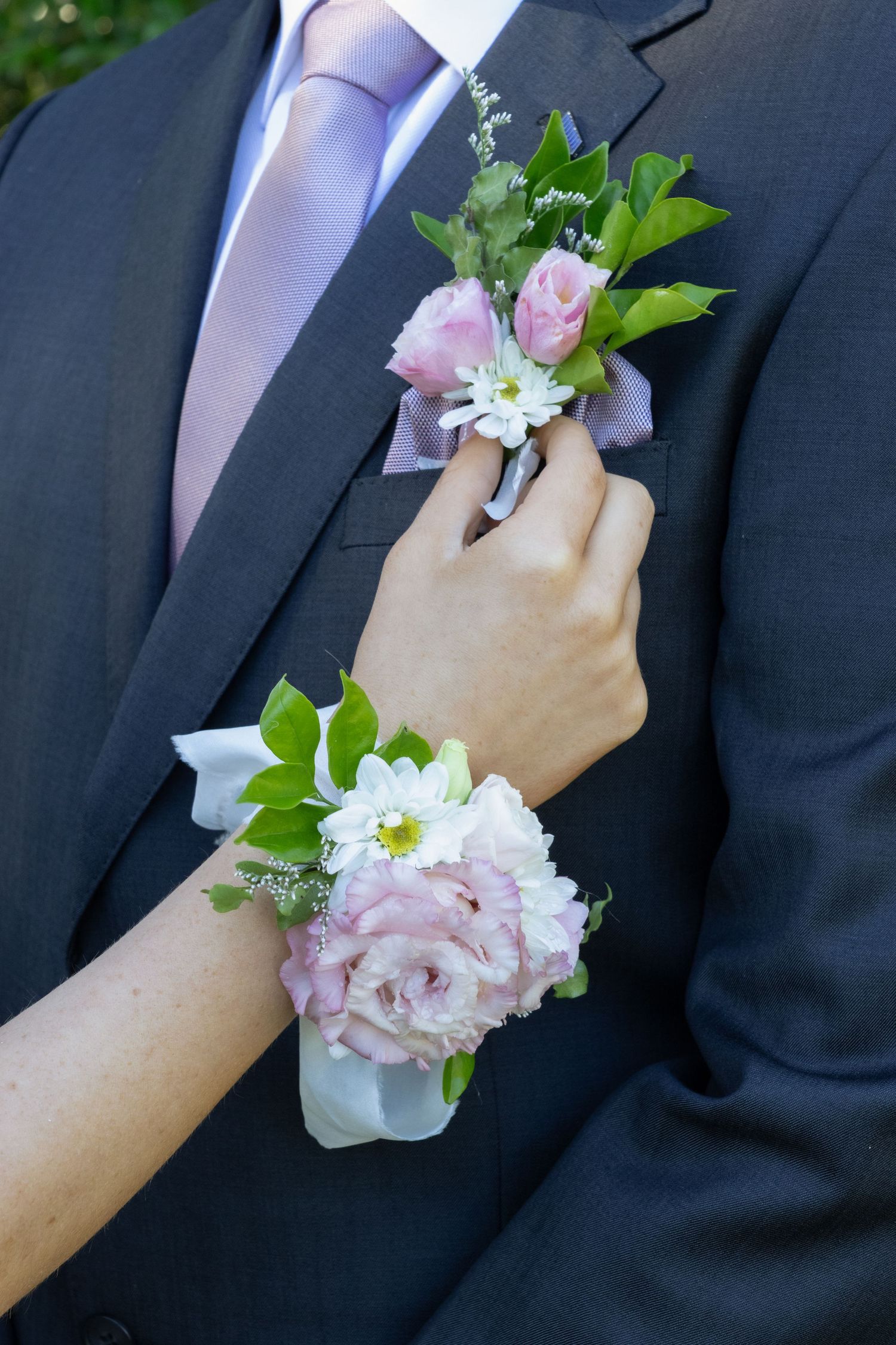 Pink floral corsage on wrist and boutonniere on dark suit lapel with lavender tie at formal event.