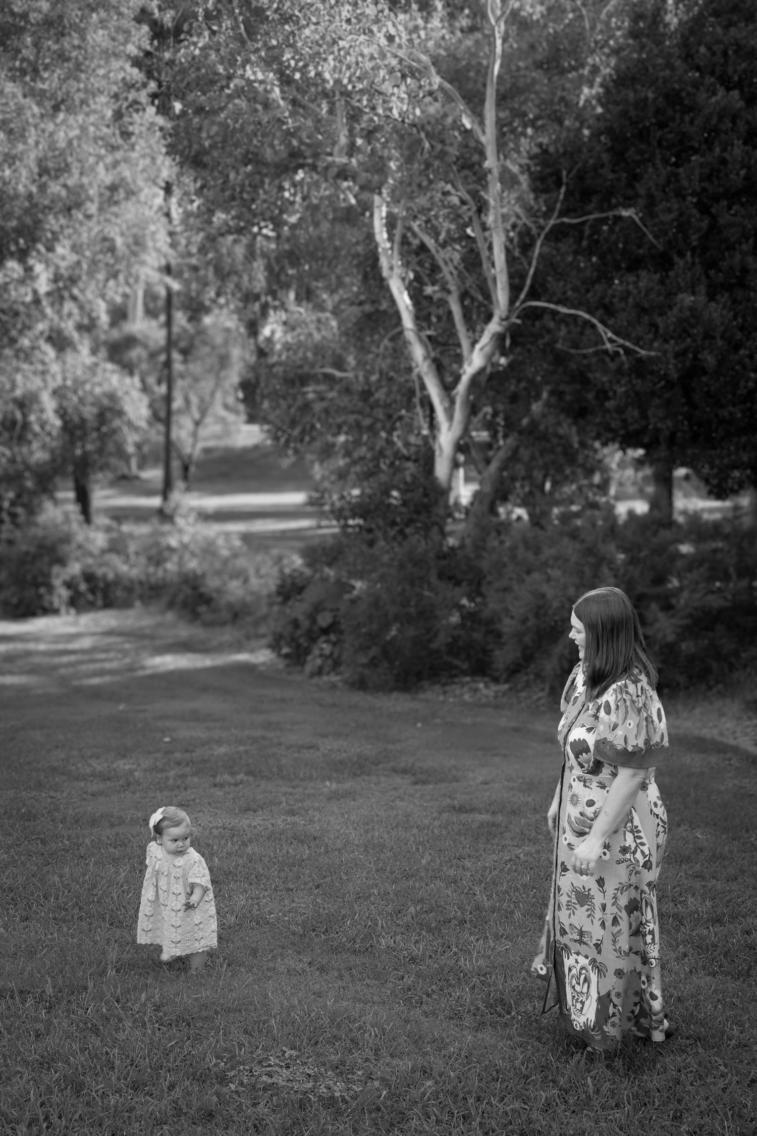 Black and white photo of a woman in a floral dress standing on grass with a toddler in a park setting.